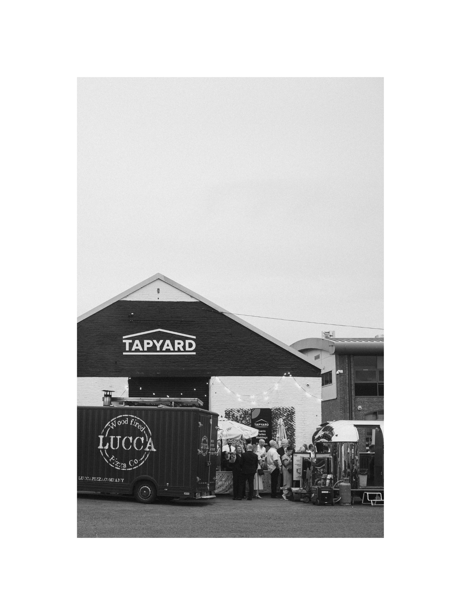 People gathering at an outdoor food market with a tapyard pizza truck and Lucca pizza stand under a large building.