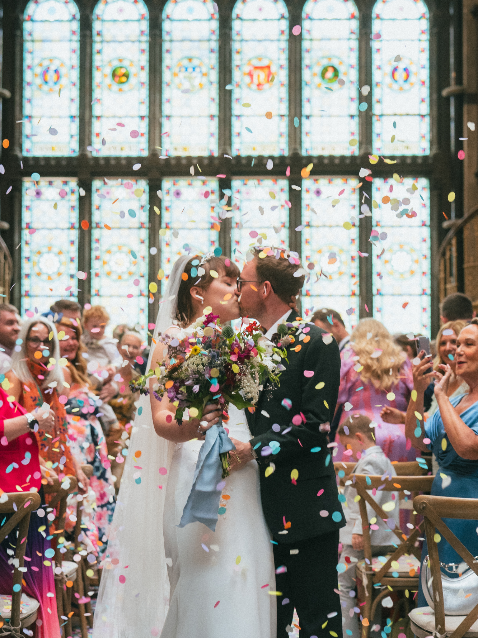 A bride and groom kiss during their wedding ceremony inside a church with stained glass windows as colorful confetti falls around them.