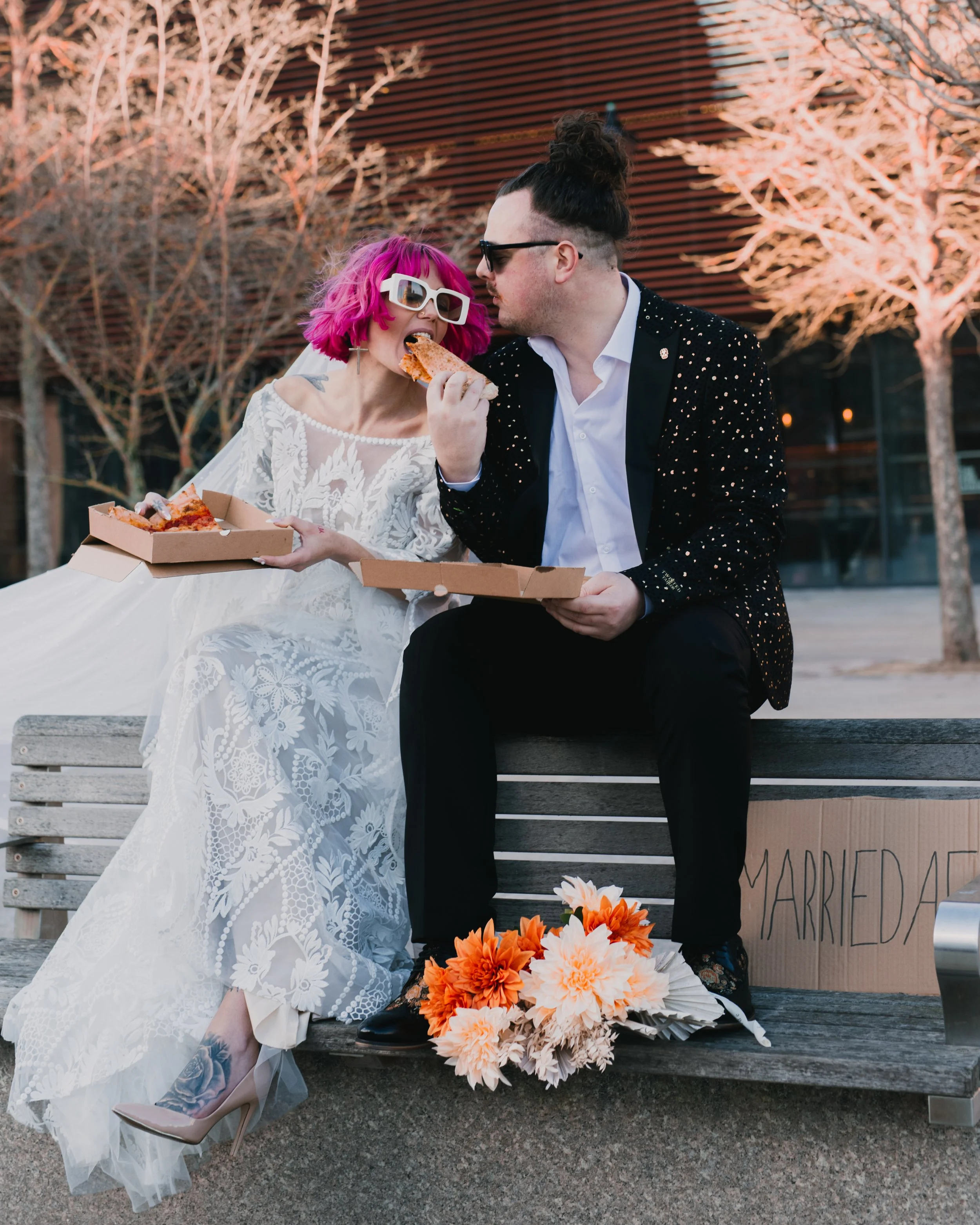 Newlywed couple in formal attire eating pizza on a bench, with flowers beside them.
