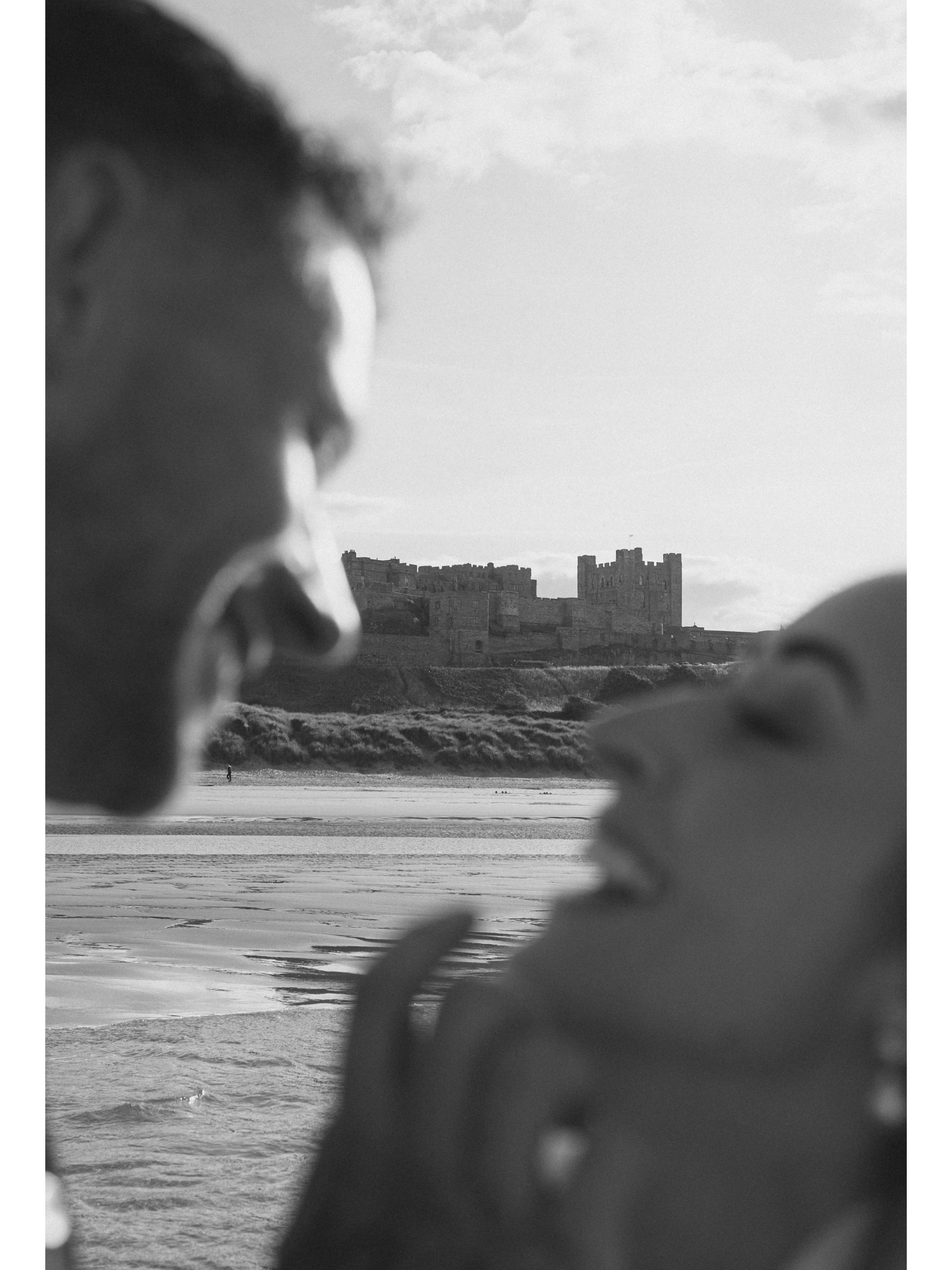A black and white photo showing a person’s face in profile on the left side, with blurred details, facing a scene of a castle in the distance across a body of water, under a cloudy sky.