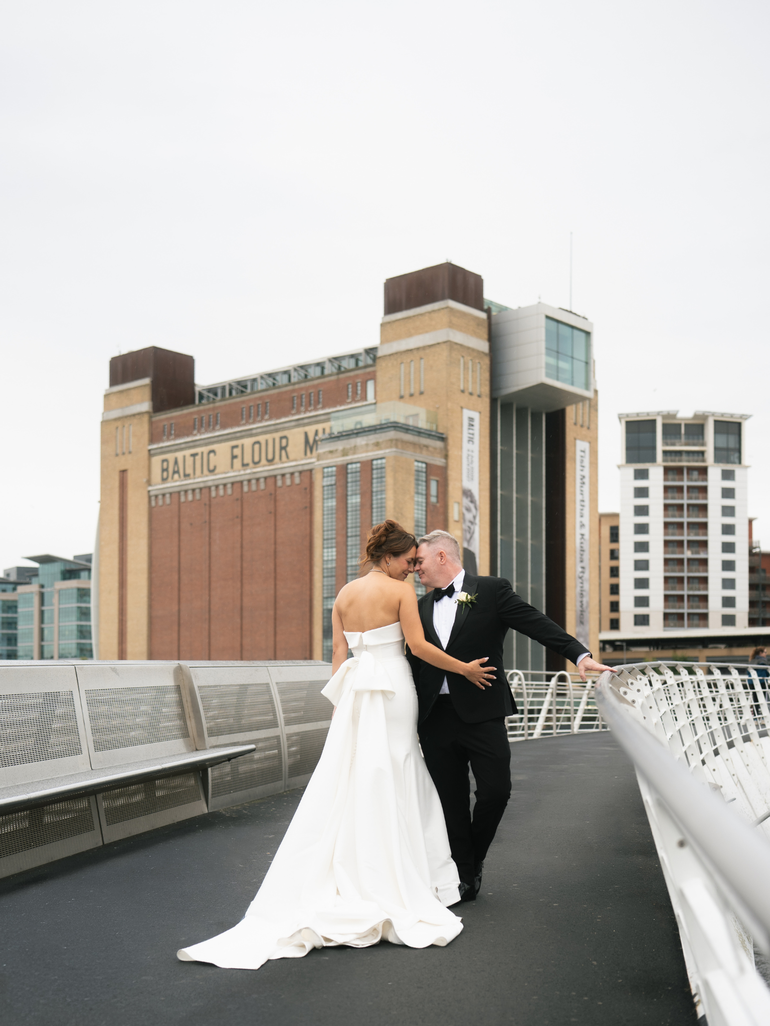 A bride and groom sharing an intimate moment on an outdoor walkway in an urban setting, with tall buildings in the background.