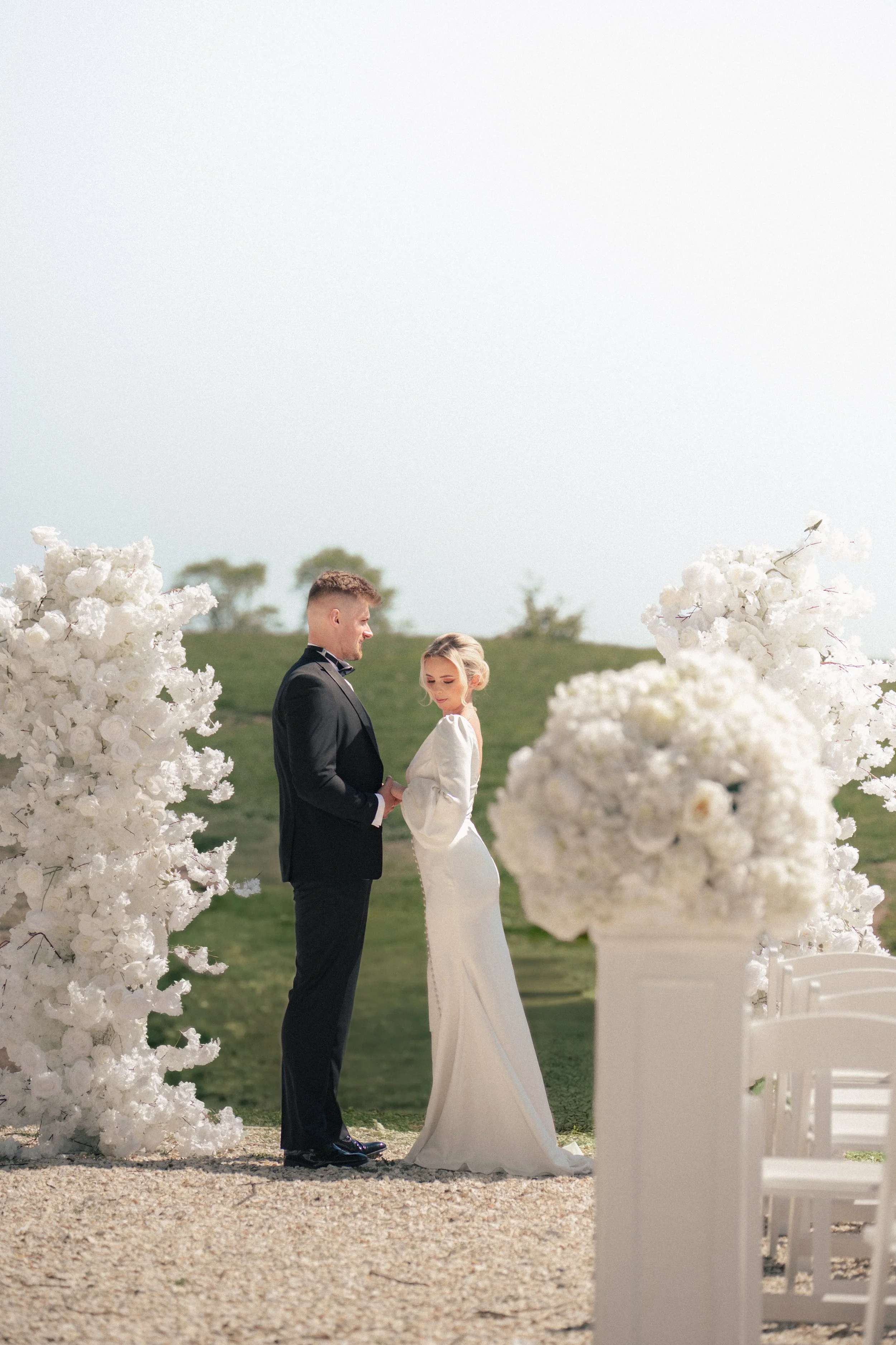 Bride and groom standing outdoors during wedding ceremony, surrounded by white floral decorations, on a gravel path with green grass and small trees in the background.