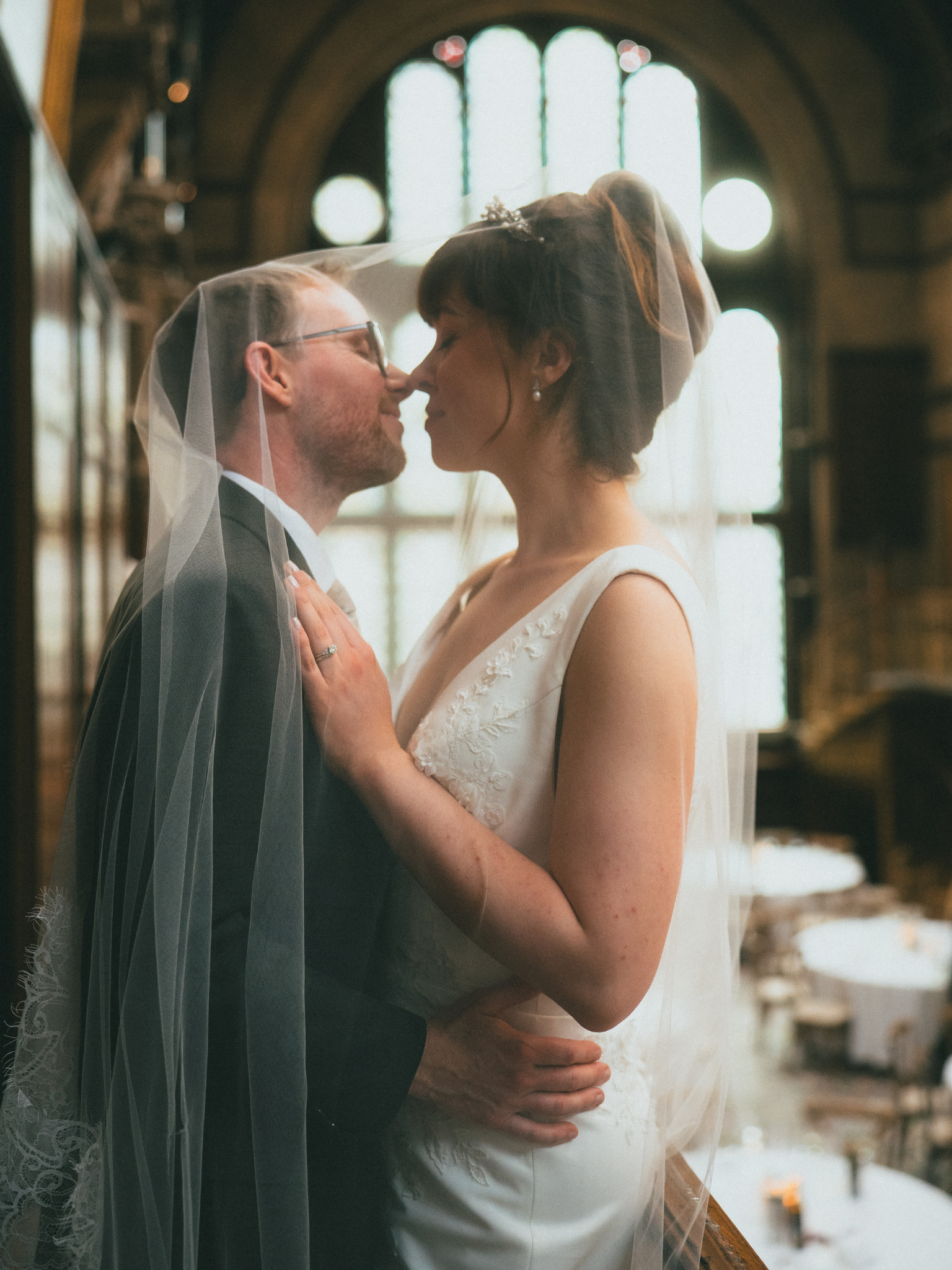 A bride and groom share a kiss inside a church. The groom wears glasses and a suit, and the bride wears a wedding dress with embroidery and a veil. They hold each other close, with the bride's hand resting on the groom's chest, and a large arched win