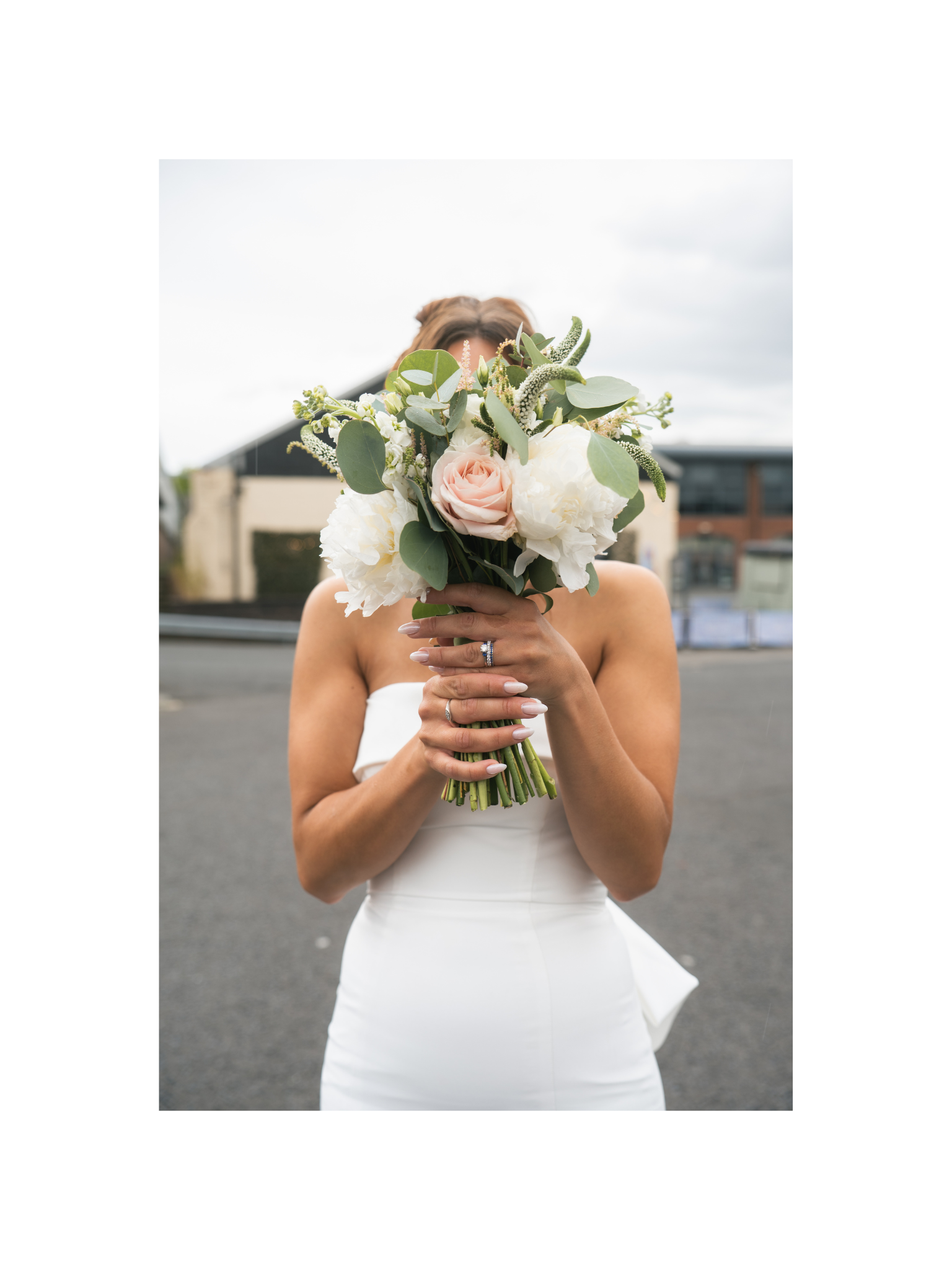 A woman in a wedding dress holding a bouquet of flowers in front of her face, standing outdoors.