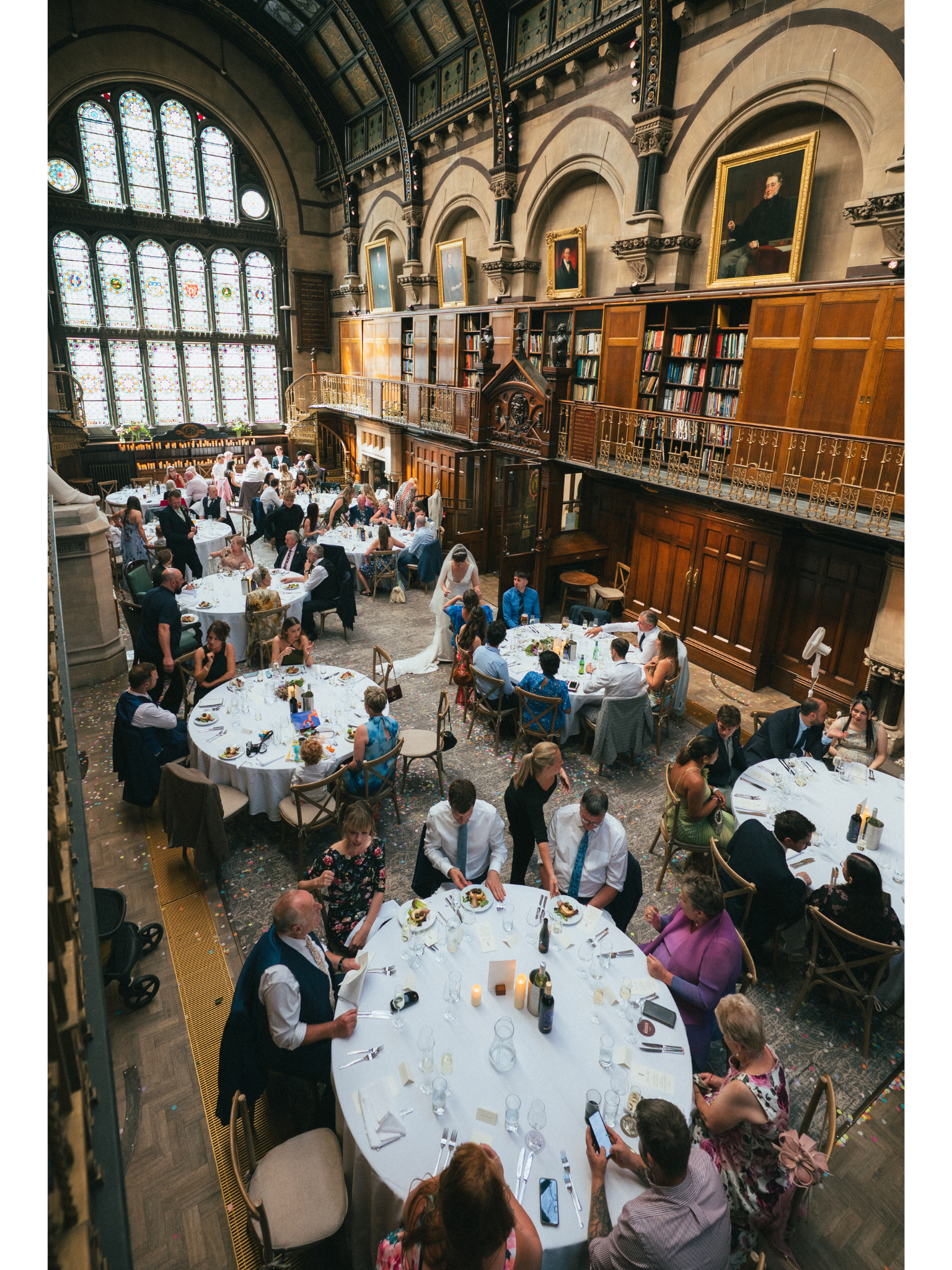 A large indoor event hall with high stained glass windows, ornate wooden wall paneling, and framed portraits. People are seated at multiple round tables with white tablecloths, eating and socializing. The room has a historic and elegant atmosphere, w