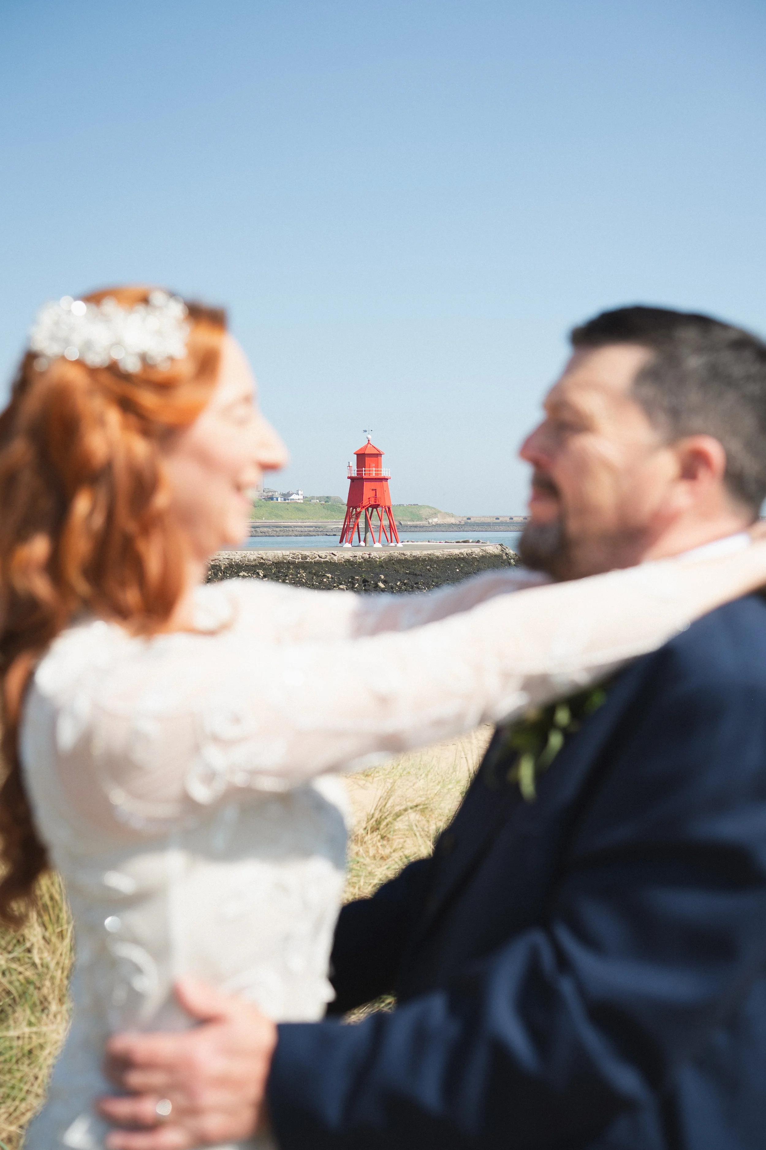 A bride and groom are smiling and embracing outdoors with a red lighthouse and blue sky in the background.