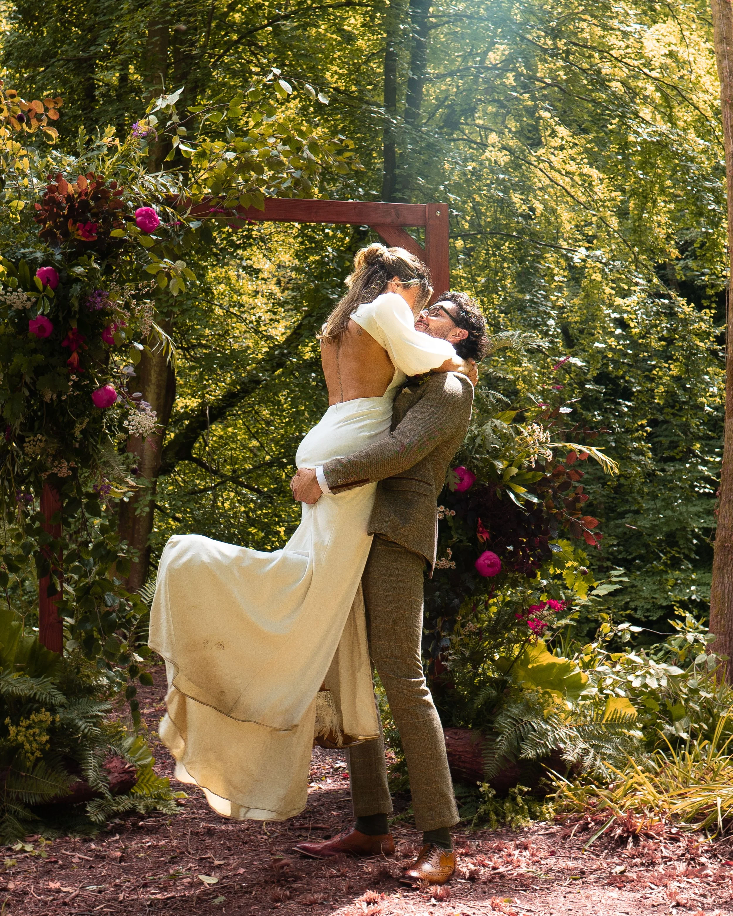 Bride and groom embrace in woodland setting with flower arch.