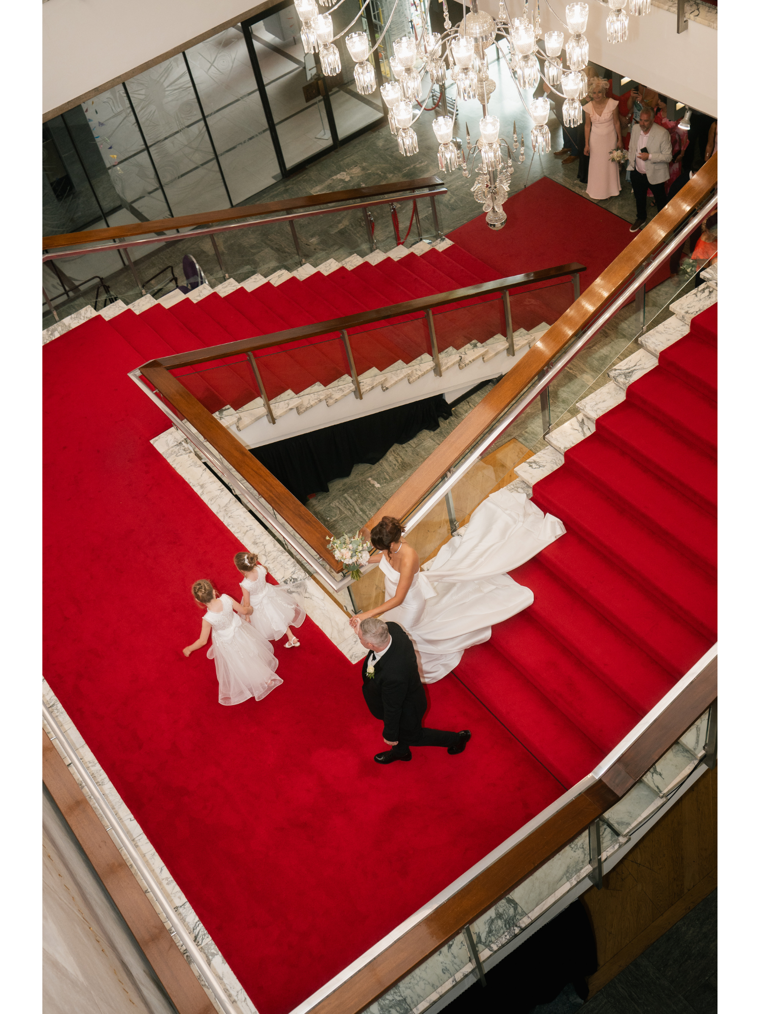 A bride in a white wedding dress walking down the stairs at a wedding reception, accompanied by three young flower girls in white dresses, with guests gathered at the bottom of the stairs.