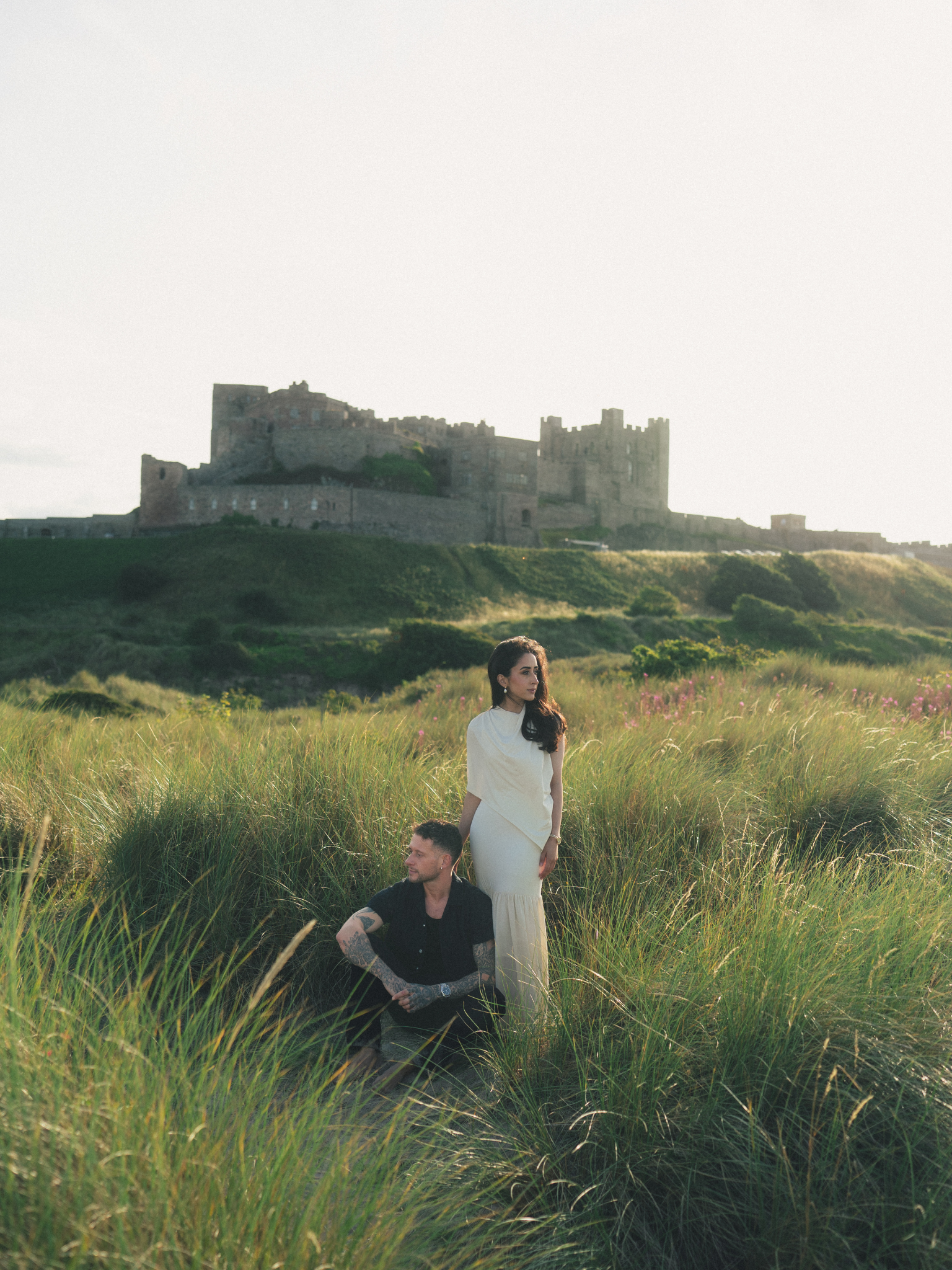 A woman in a white dress and a man with tattoos sitting in a field of tall grass with a castle on a hill in the background.