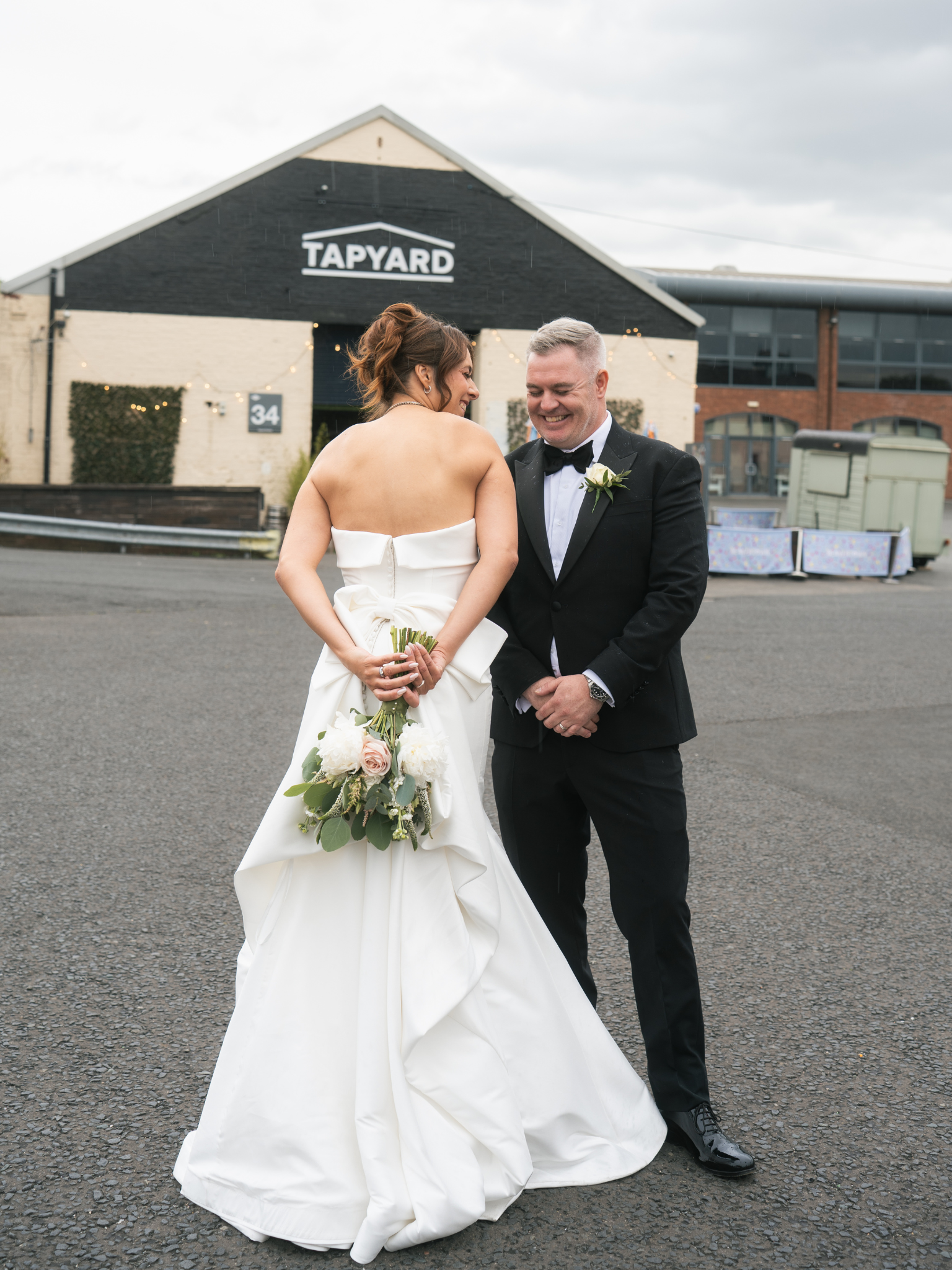 A bride in a white wedding dress holding a bouquet of roses and greenery, smiling at a groom in a black tuxedo with a white boutonniere, outside on a cloudy day in front of a building with a sign that says "TAPYARD."