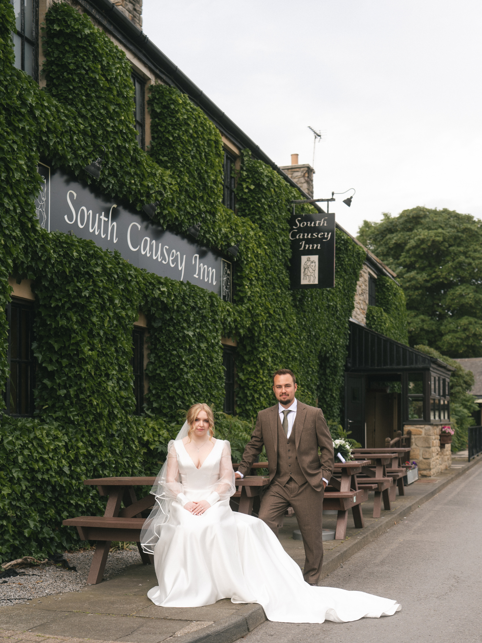 A bride and groom pose outside in front of a green ivy-covered building on a cloudy day. The bride wears a white wedding gown with long sleeves and a veil, seated at a picnic table. The groom stands beside her, dressed in a brown suit, white shirt, a
