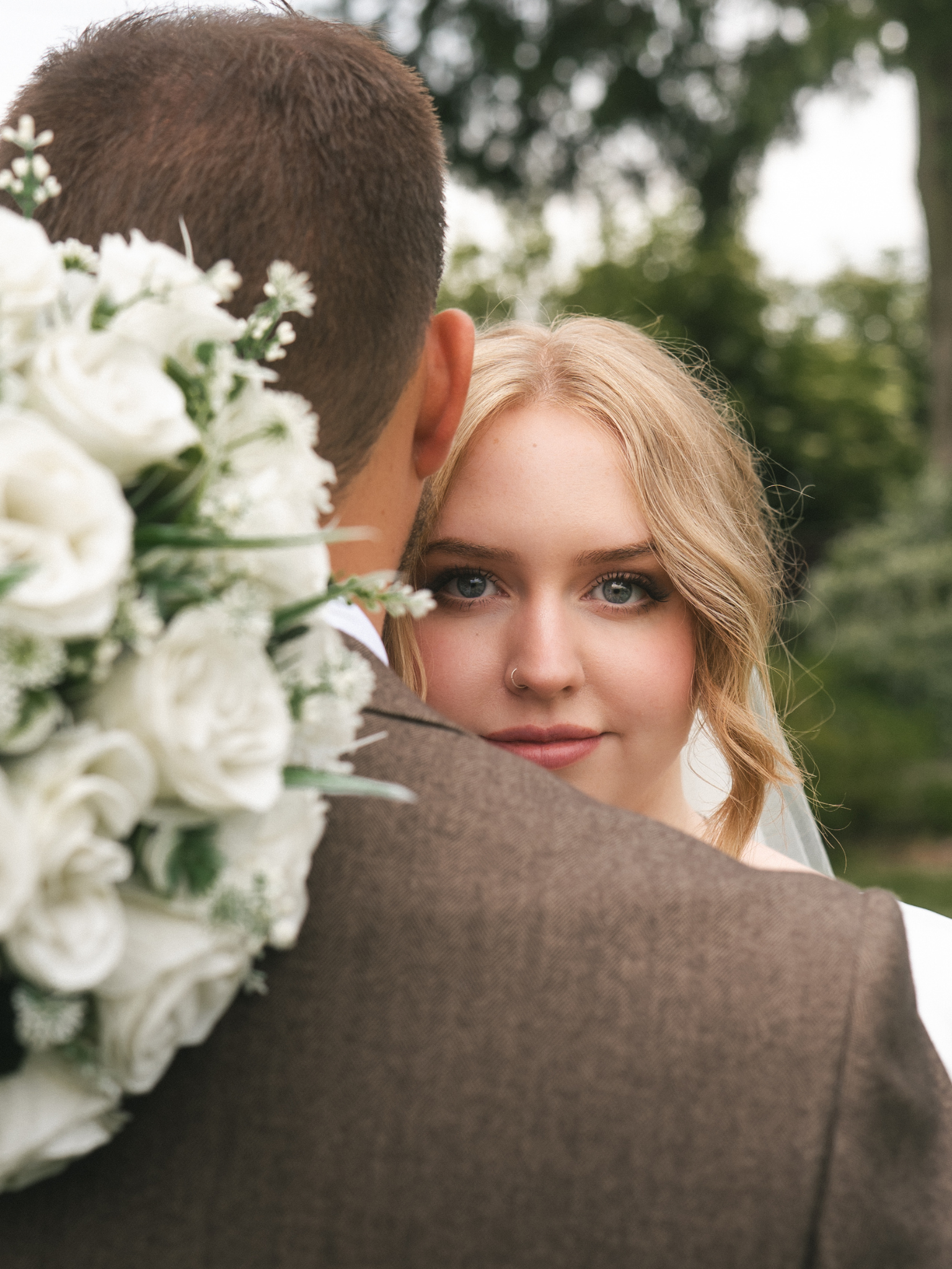 A bride with blonde hair and blue eyes looking at the camera is being embraced by a groom with short brown hair, outdoors with trees in the background, with a bouquet of white flowers partially visible.