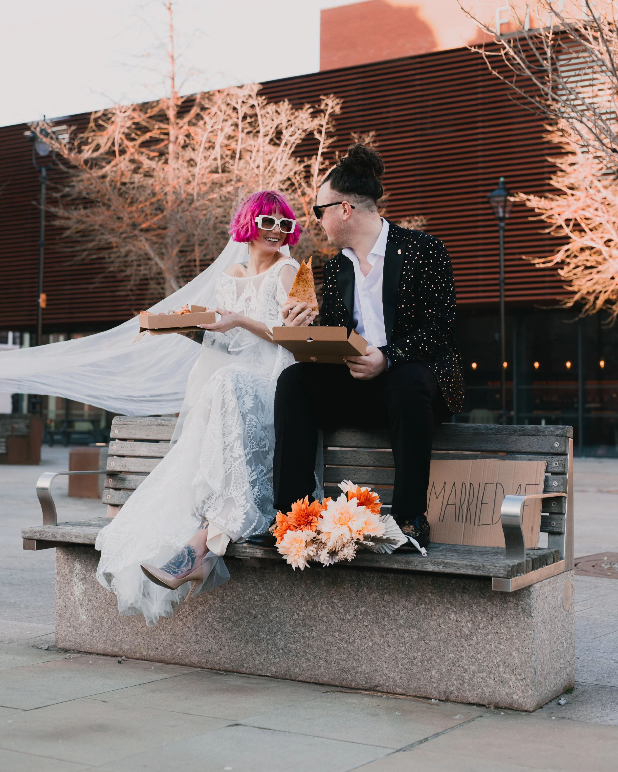 Bride and groom sitting on a bench, bride in a white dress with pink hair and sunglasses, groom in a black suit with a top knot hairstyle, eating pizza. Bouquet of flowers and a 'Just Married' sign beside them.