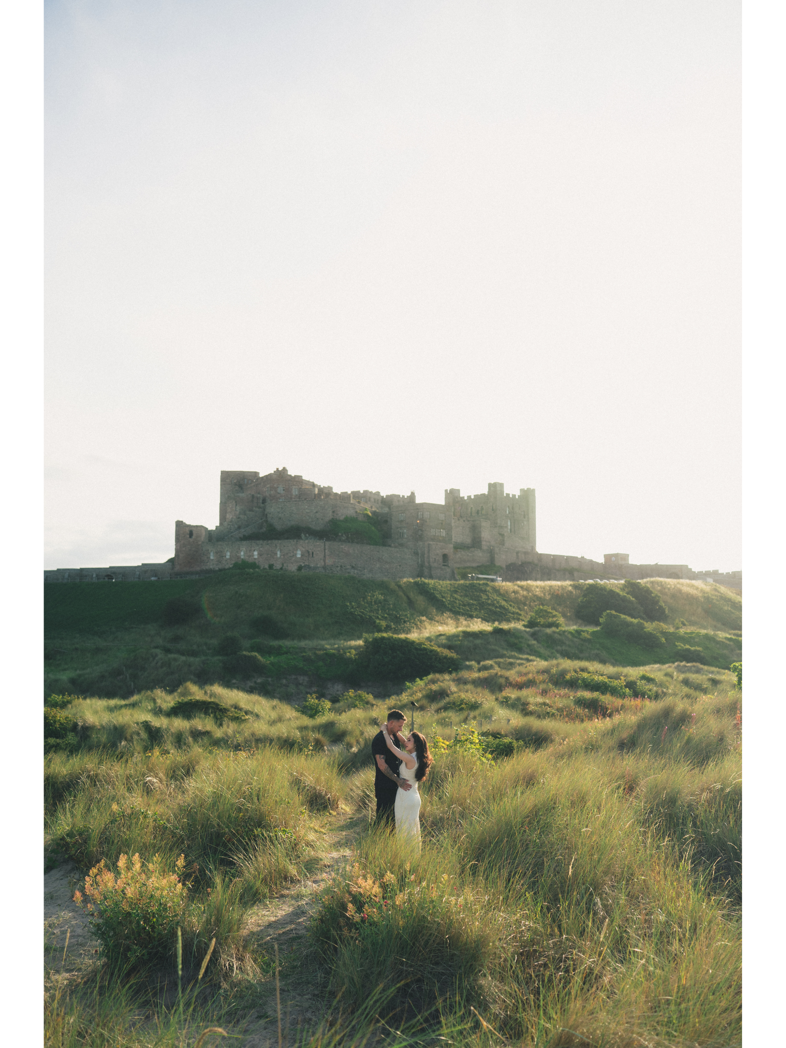 A couple stands in a lush grassy field with a castle on a hill in the background. The man and woman are holding each other's hands, smiling, with the sun setting behind the castle.