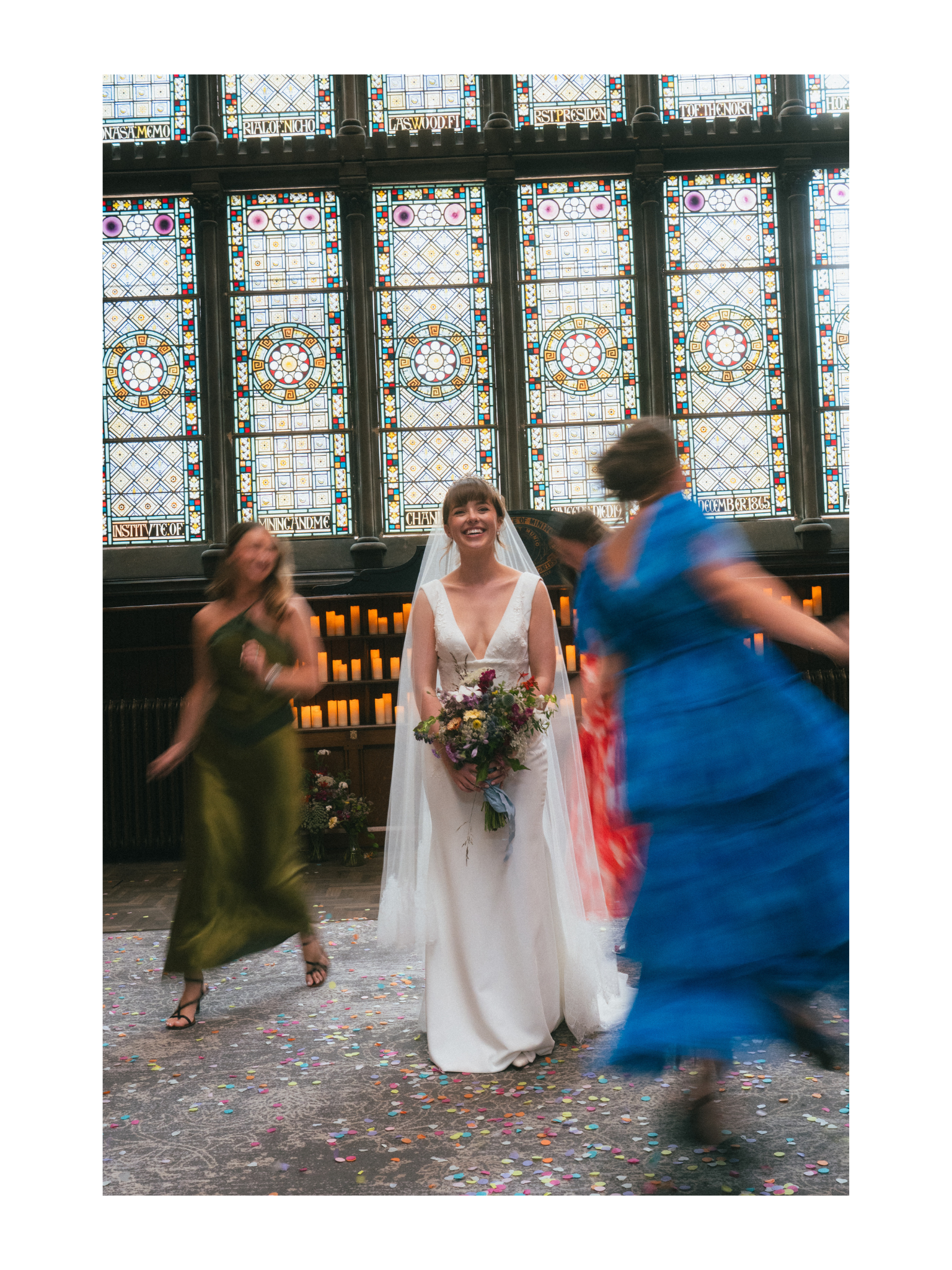A woman in a white wedding dress smiling while holding a bouquet of flowers, surrounded by dancing guests and colorful confetti on the floor inside a venue with stained glass windows in the background.