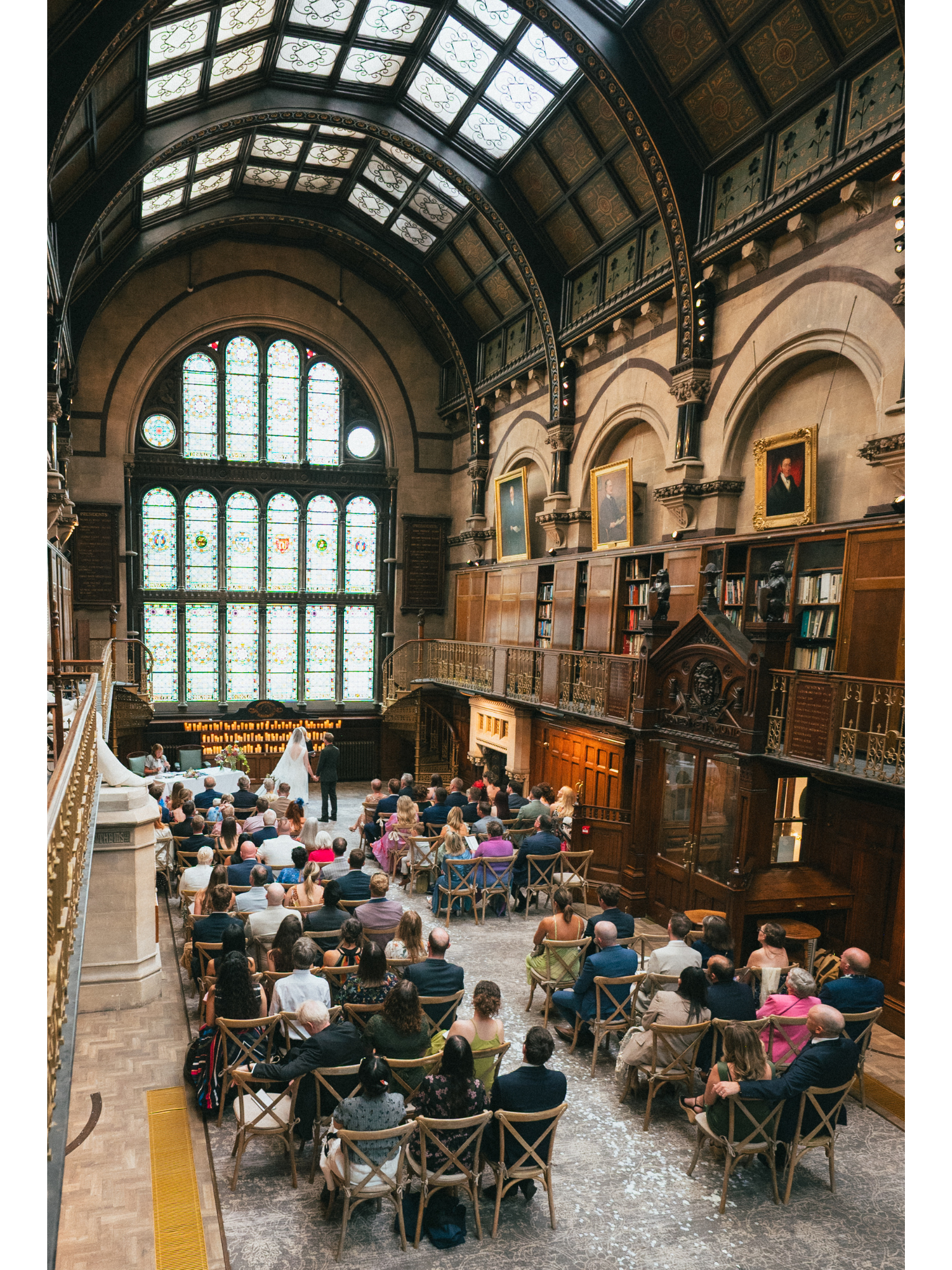 A wedding ceremony inside a historic building with large stained glass windows and a high ornate ceiling. Guests are seated facing the couple and officiant, with portraits hanging on the walls and warm lighting.