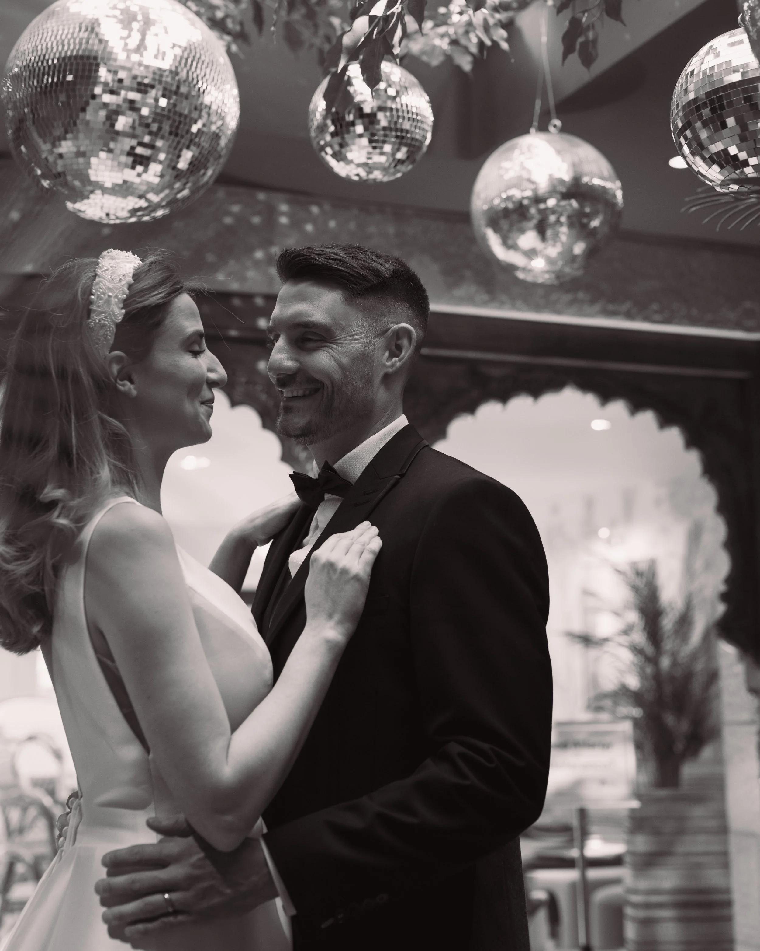Black and white photo of a couple smiling and embracing at a wedding, with disco balls hanging above.