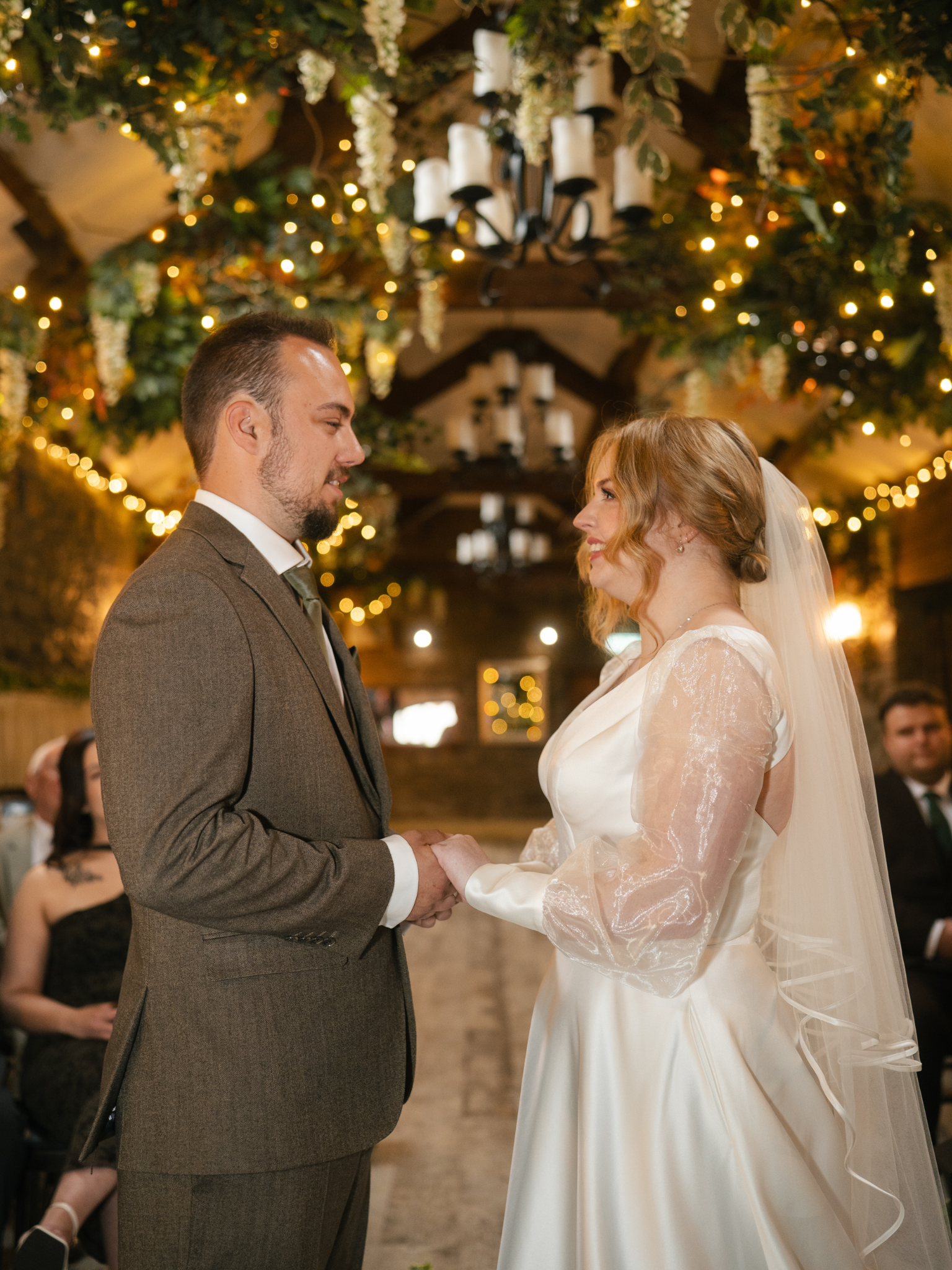 A bride and groom holding hands during their wedding ceremony, with a decorated ceiling featuring hanging flowers and string lights.