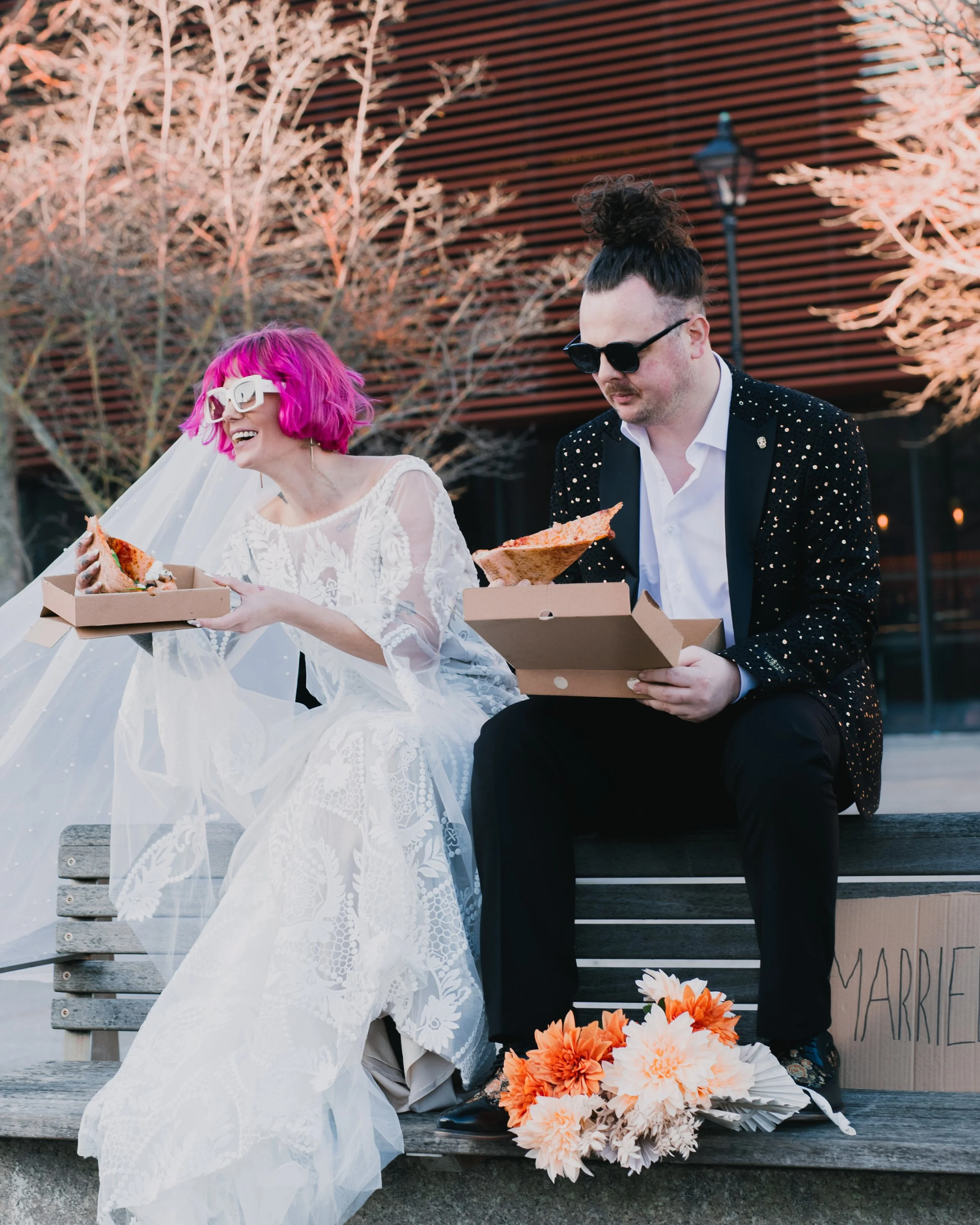 Bride and groom eating pizza on a bench, wedding attire, bouquet on ground, casual setting.