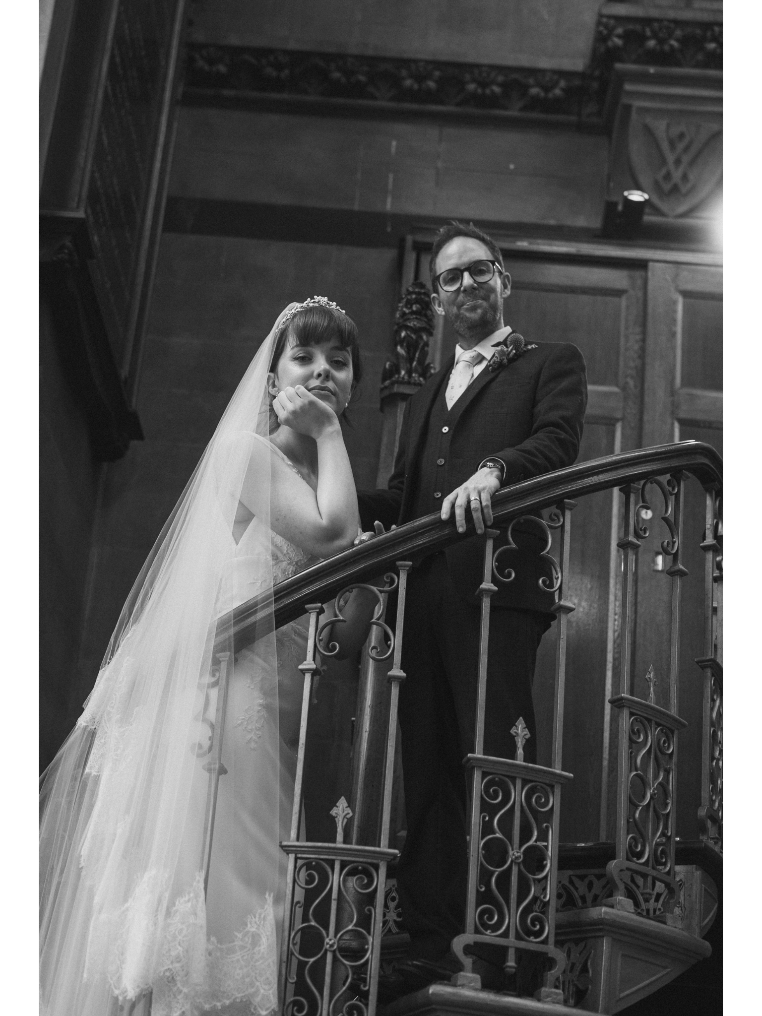 A bride and groom standing on a staircase with ornate railing, inside a wooden-paneled building with decorative details.