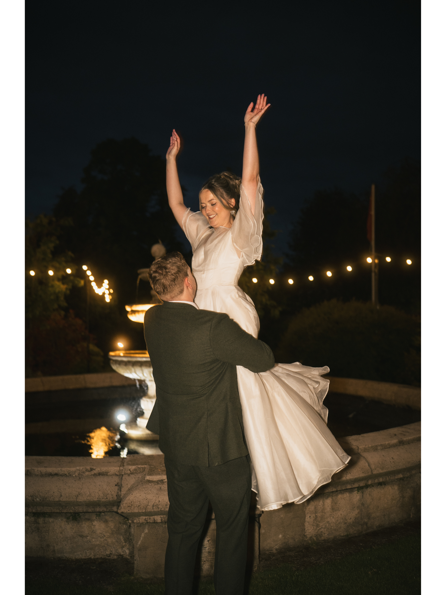 A groom lifting and holding a smiling bride with her arms raised at night near a fountain with string lights in the background.