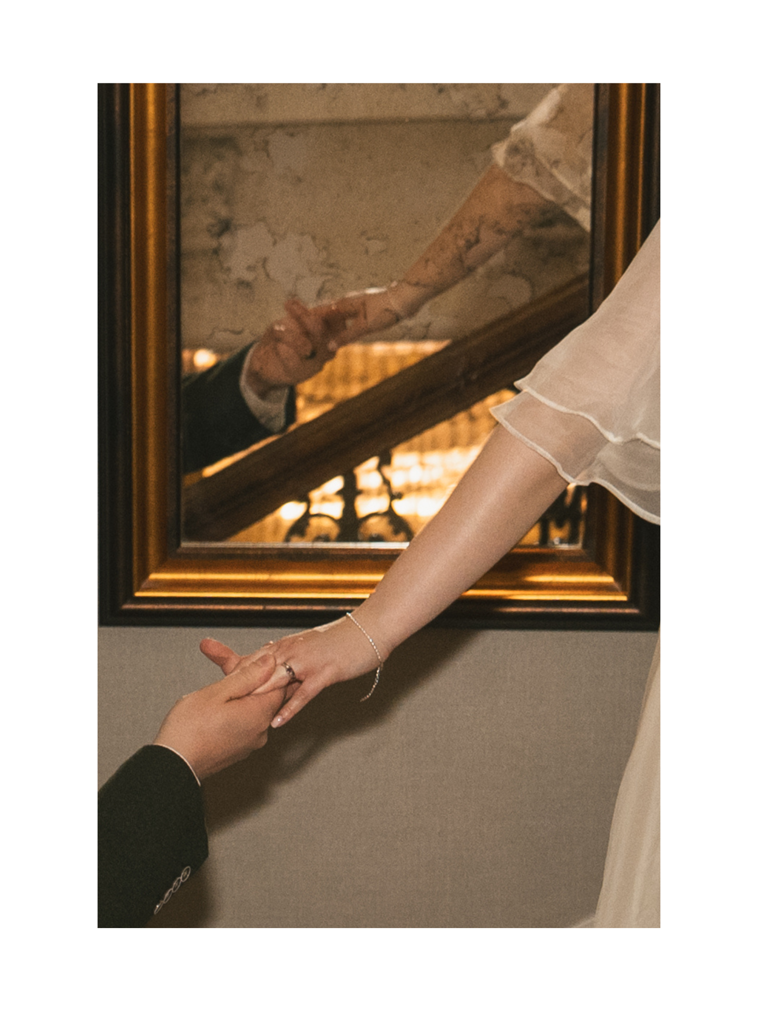 A close-up photo of two people holding hands, with a mirror reflecting the scene. One person wears a white dress or shirt and a bracelet, while the other wears a dark suit, and the setting appears formal or romantic.