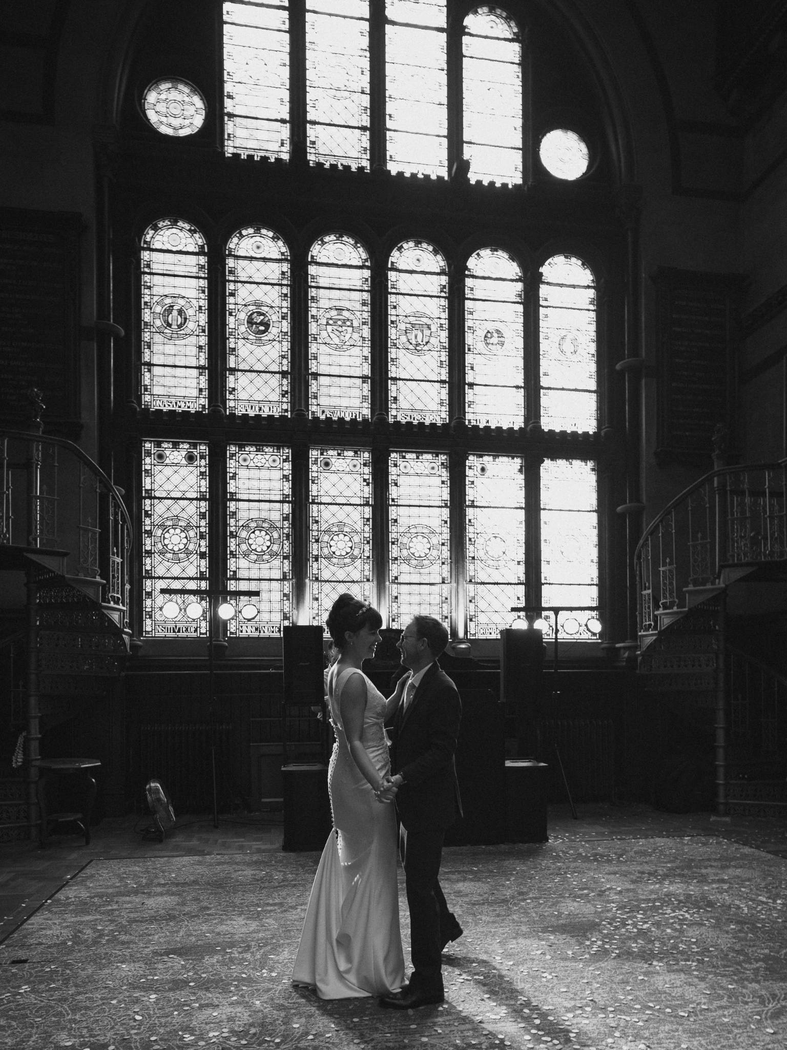 A couple in wedding attire standing close together and smiling at each other inside a church with large stained glass windows and a high vaulted ceiling.