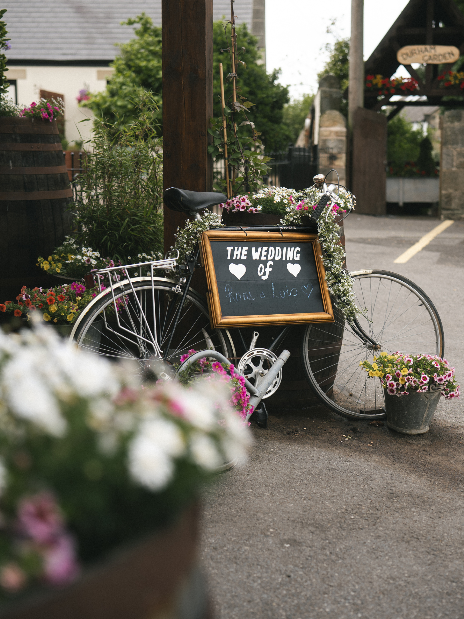 Decorative bicycle with flowers and a sign for a wedding, placed outdoors near a building and greenery.