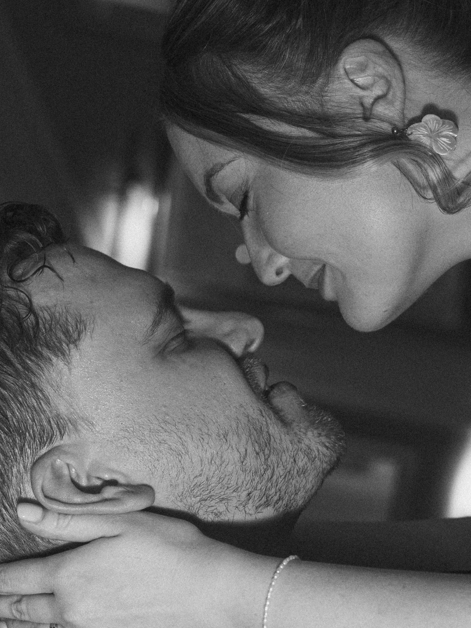 A close-up black-and-white photo of a woman with earrings, with her eyes closed and smiling, resting her head against a man's head in an intimate moment.