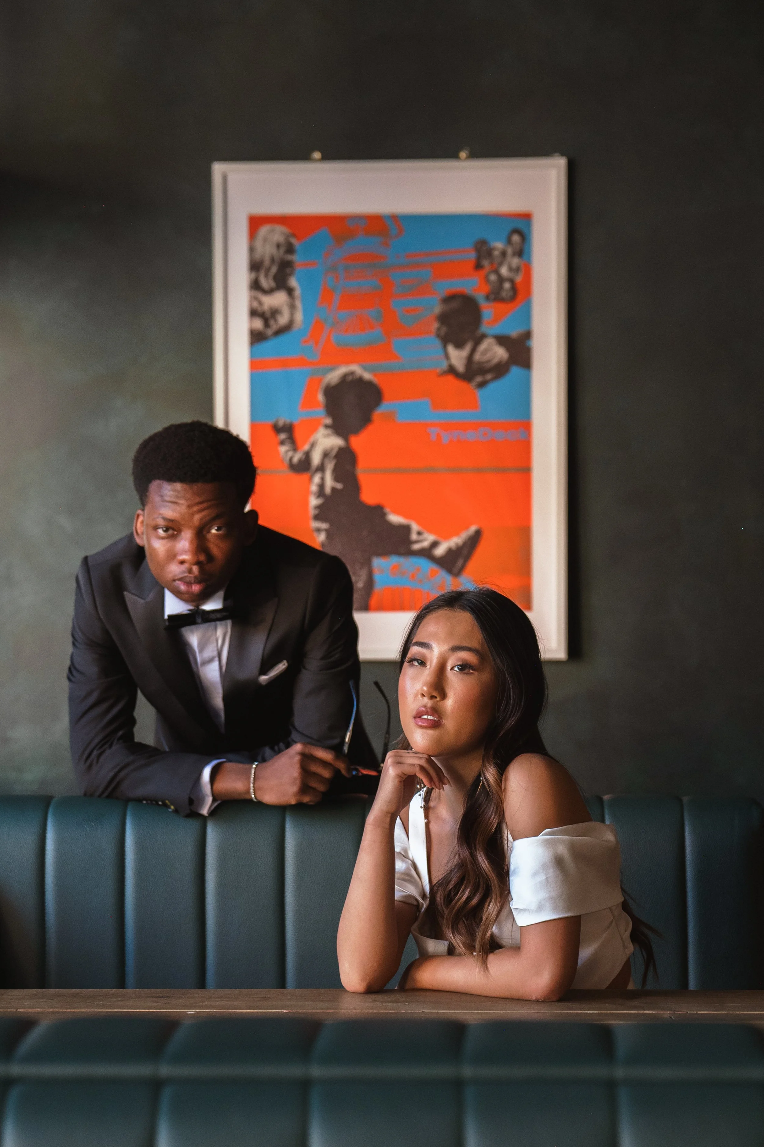 Two people in a stylish setting, one man standing, one woman seated, with colorful art in the background.
