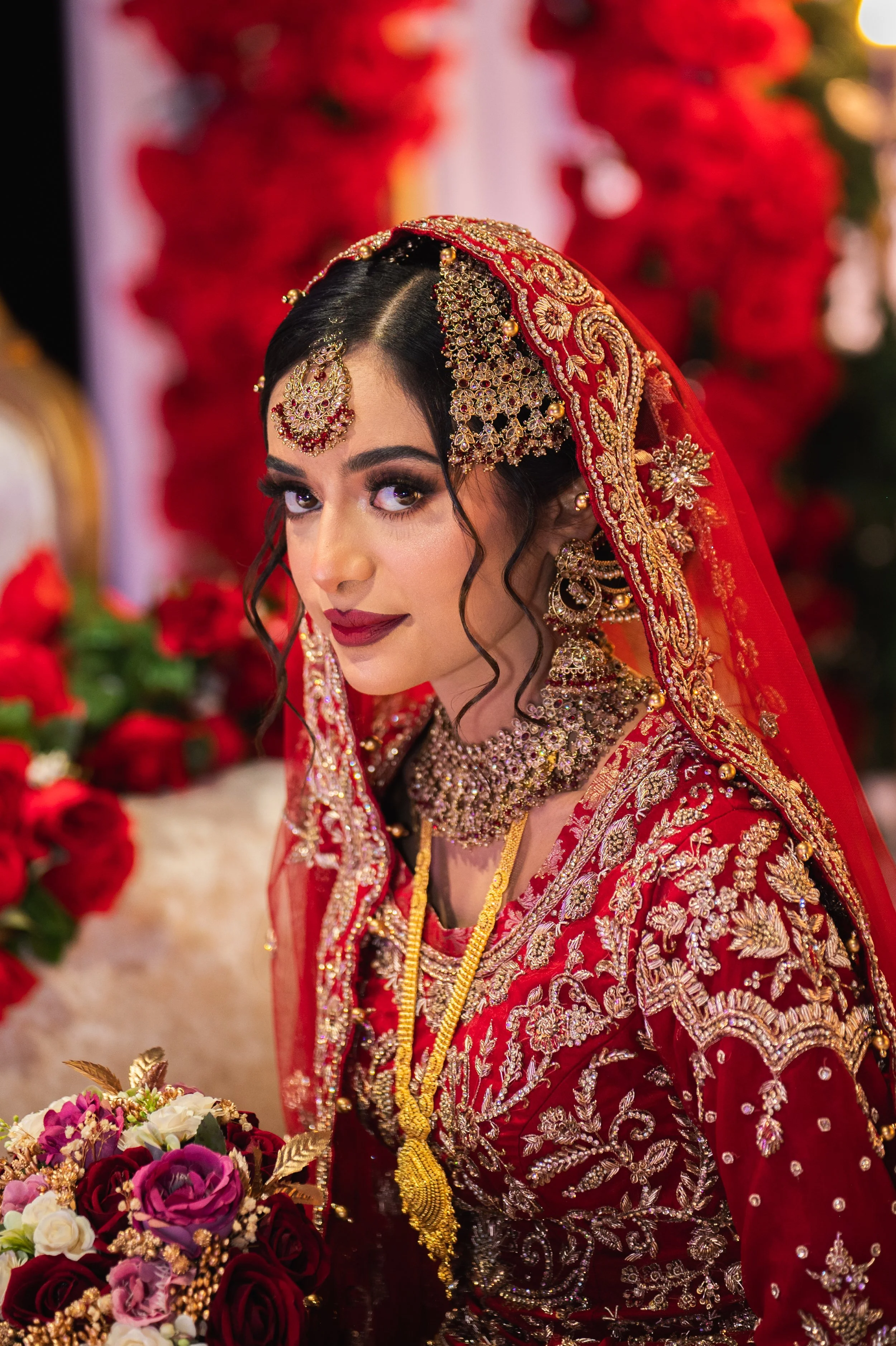 Bride in traditional red and gold bridal attire with intricate embroidery, holding a bouquet of flowers, set against a floral backdrop.