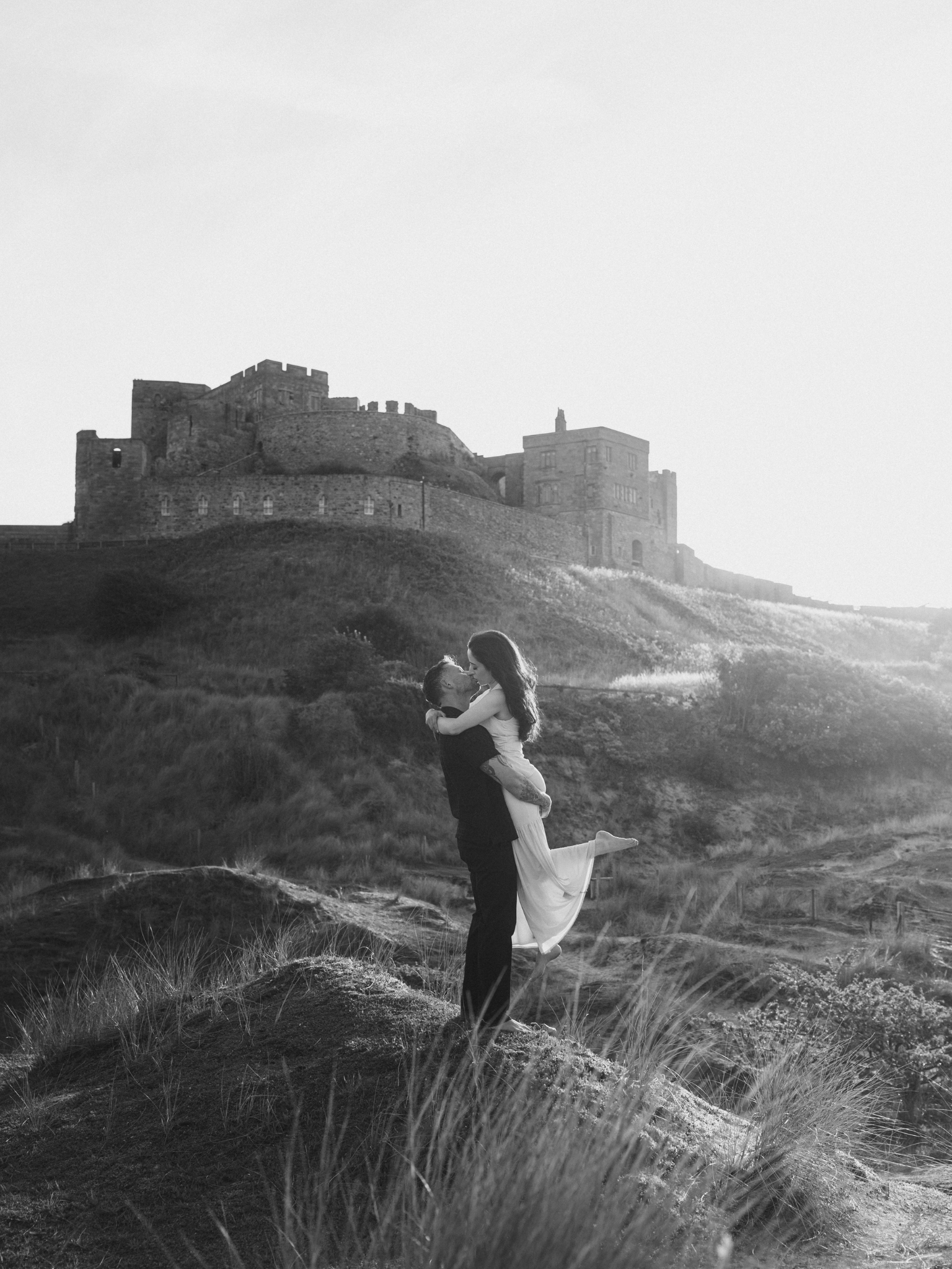 A black and white photo of a couple, with the man lifting the woman in his arms, near a grassy hill with a large castle or fortress in the background, under a clear sky.