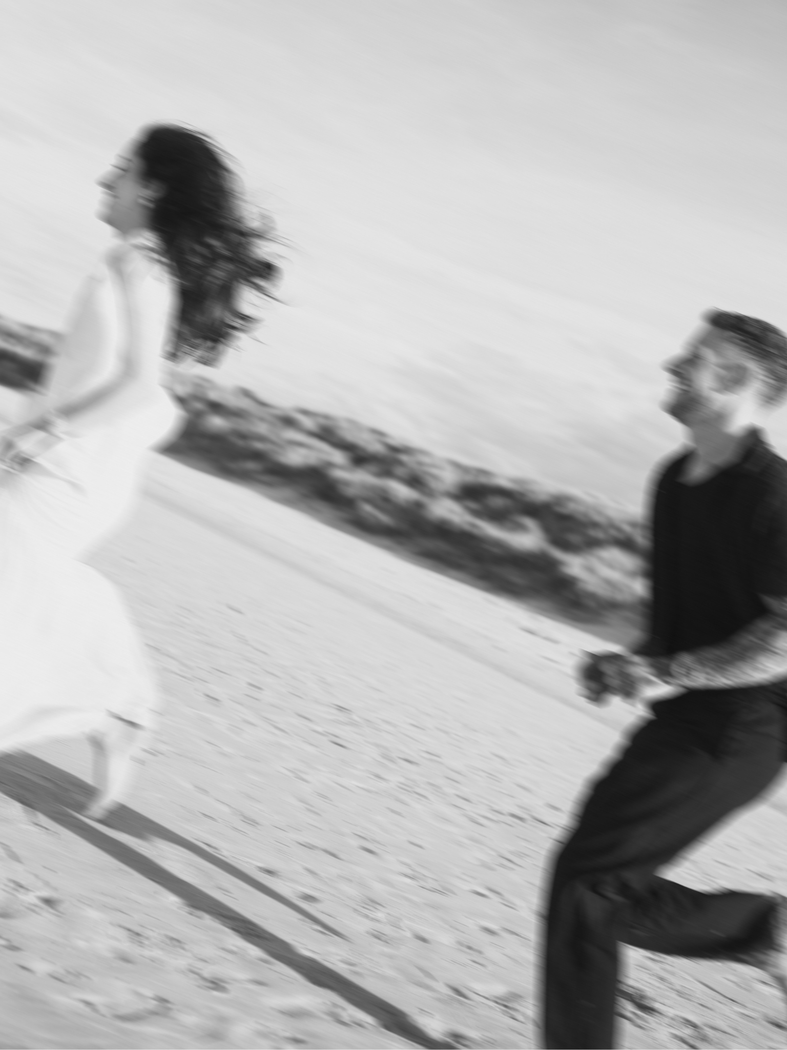 Black and white photo of a woman with long hair running or walking on snow, smiling, with a man in the background looking at her, both outdoors.