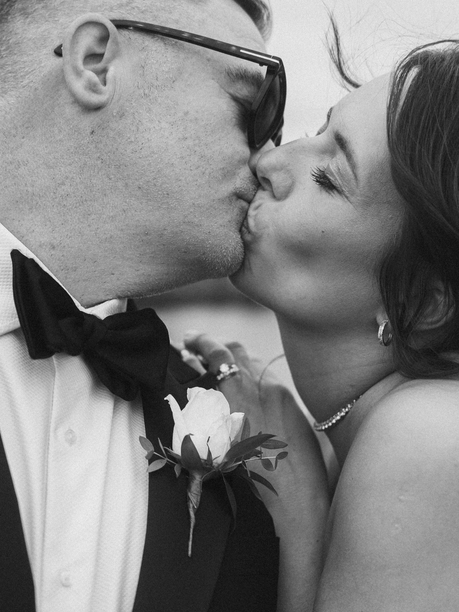 A black and white photograph of a couple sharing a kiss, dressed in formal wedding attire. The man is wearing glasses, a tuxedo with a bow tie, and a boutonniere with white flowers. The woman has earrings, a necklace, and a wedding dress with a veil,