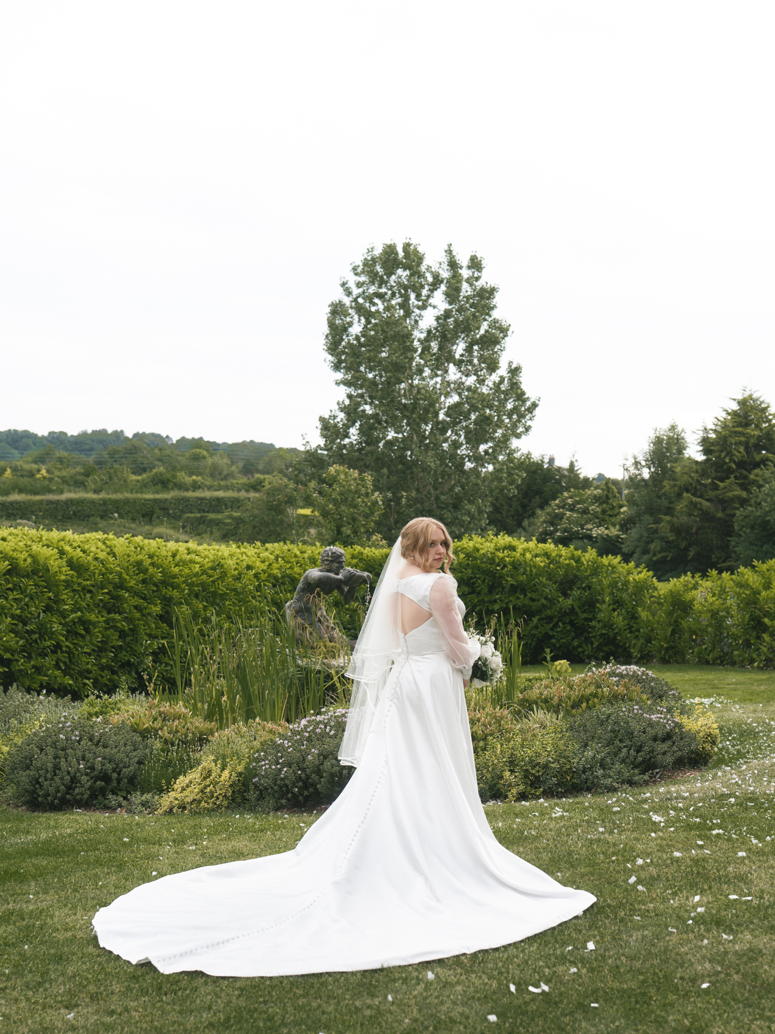 A bride stands in a garden with greenery and flowers, wearing a white wedding dress and veil, holding a bouquet, with a sculpture in the background and trees and hills in the distance.