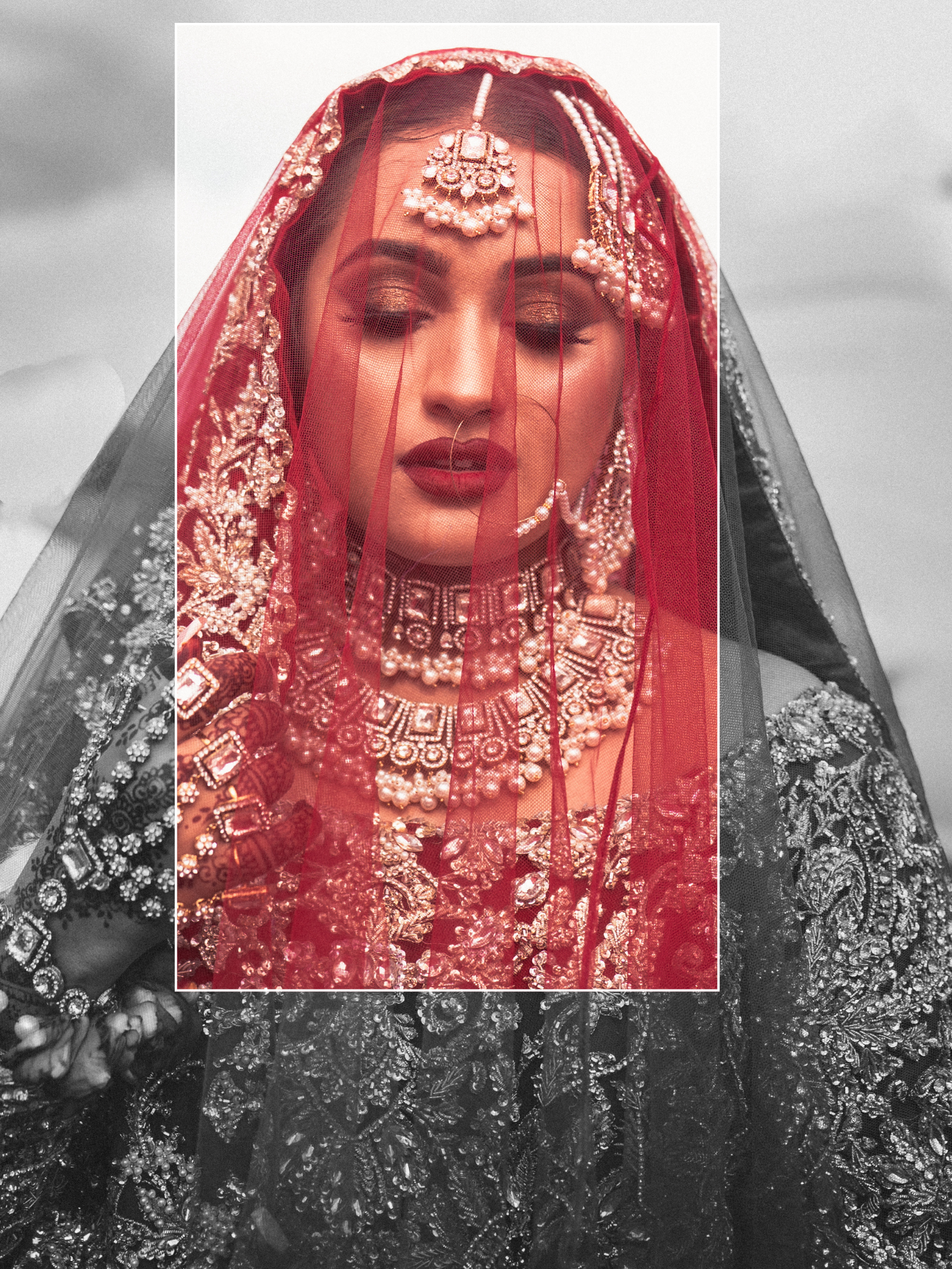 A bride dressed in traditional South Asian bridal attire with intricate jewellery, a red veil, and ornate clothing, with a background of a cloudy sky.