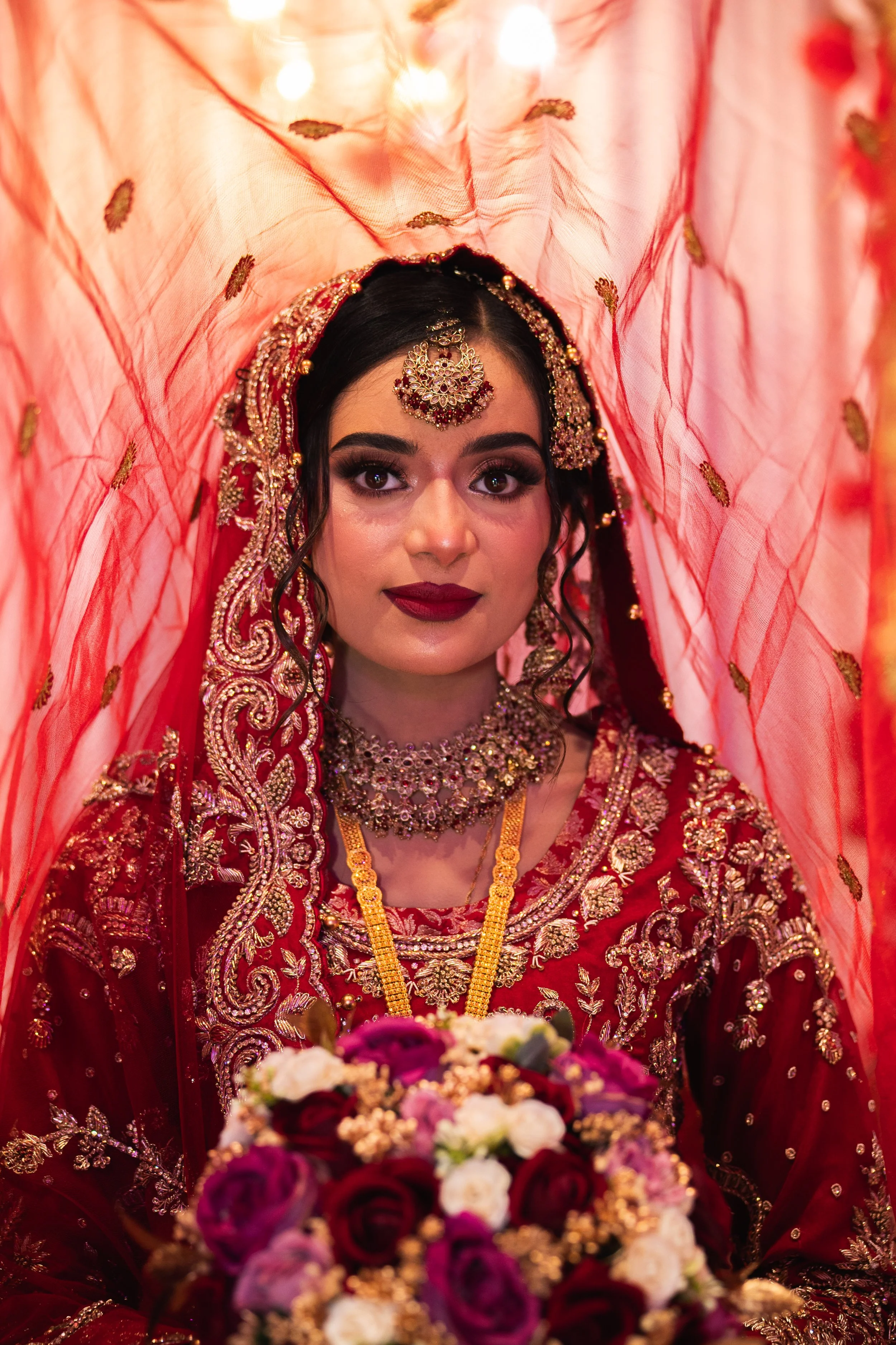 A woman in traditional bridal attire with a red veil and intricate gold embroidery, holding a bouquet of flowers.