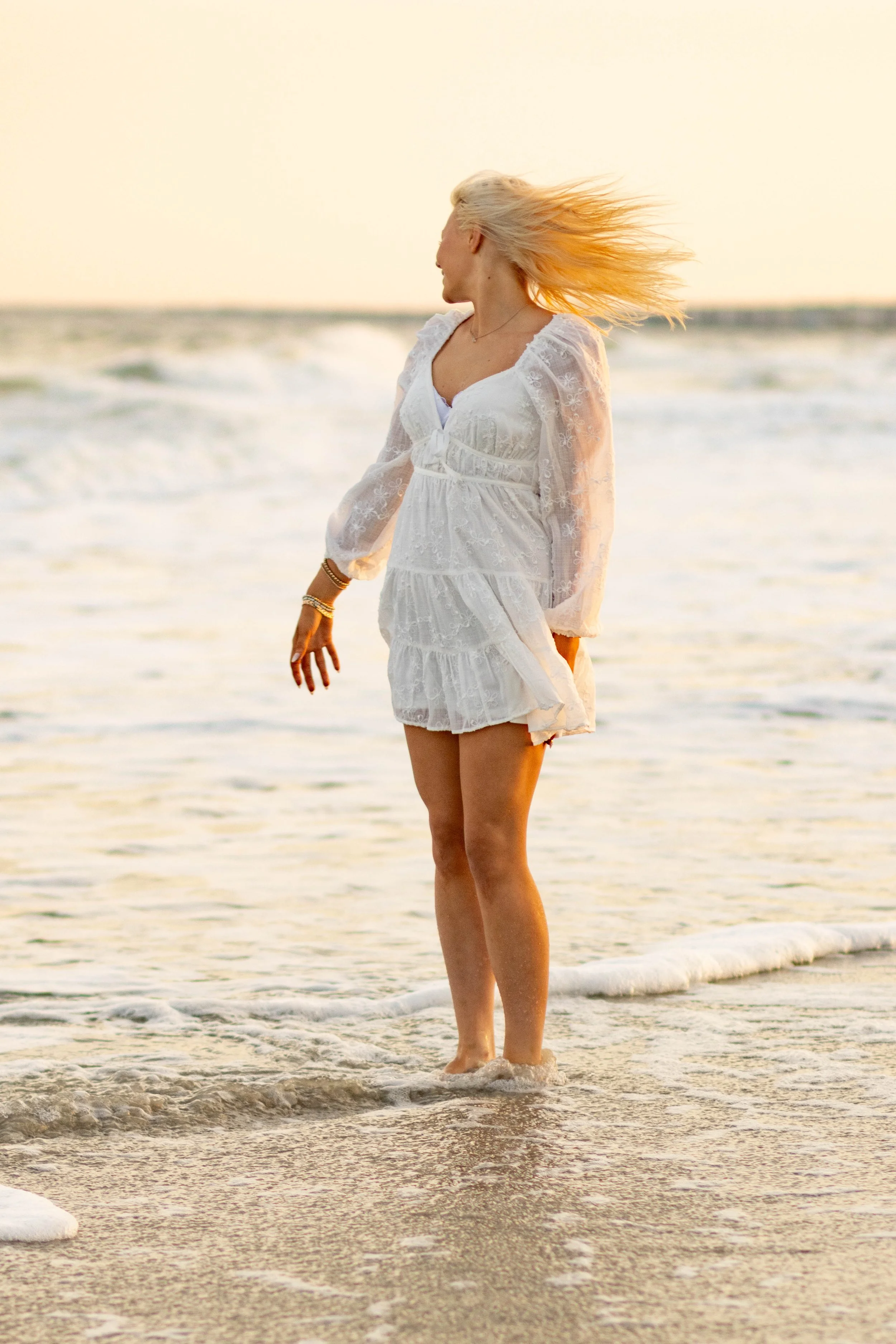 High school senior in Myrtle Beach wearing cap and gown during golden hour beach photo session, splashing in the water and smiling, taken by Myrtle Beach family and senior photographer.