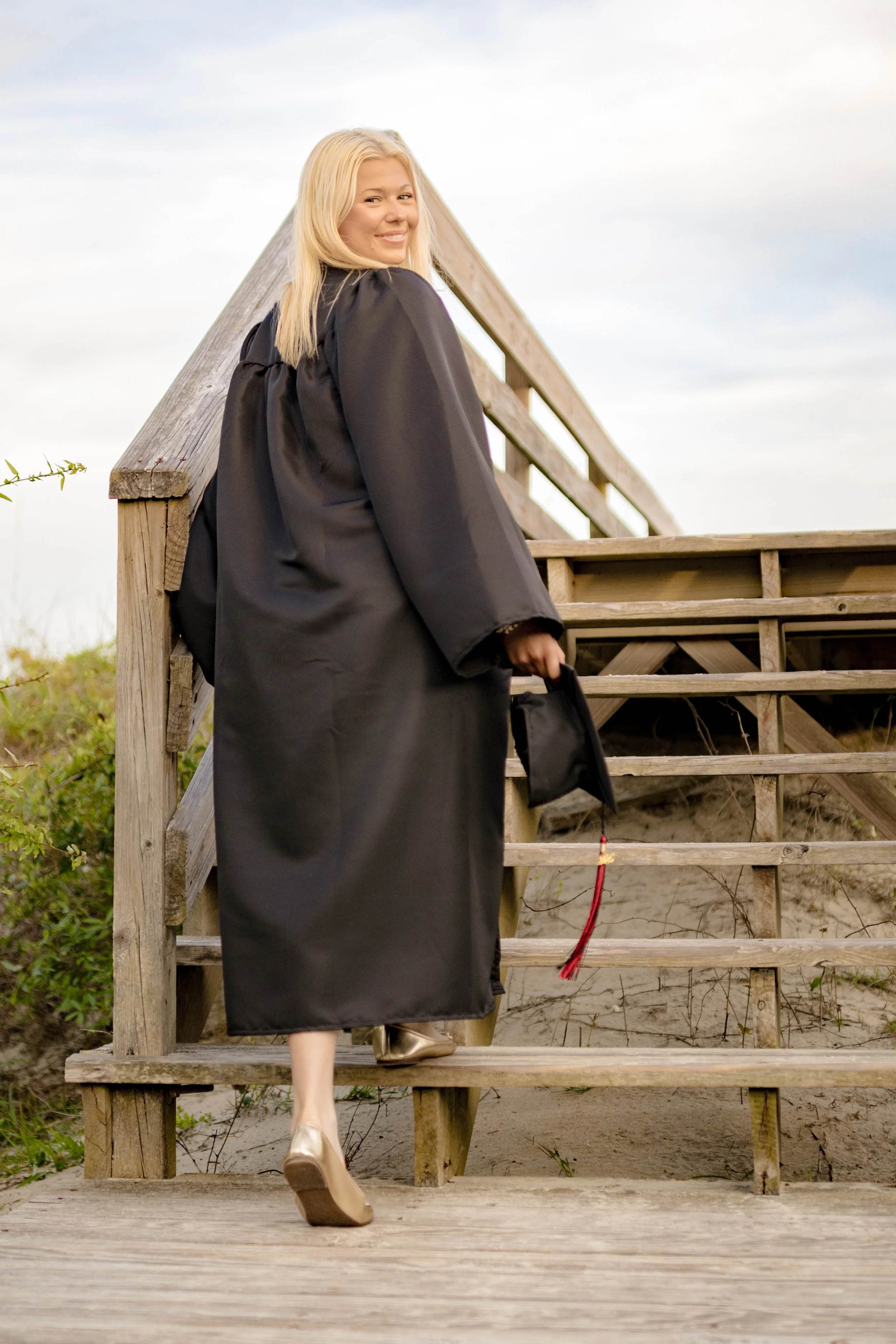 High school senior in Myrtle Beach wearing cap and gown during golden hour beach photo session, splashing in the water and smiling, taken by Myrtle Beach family and senior photographer.
