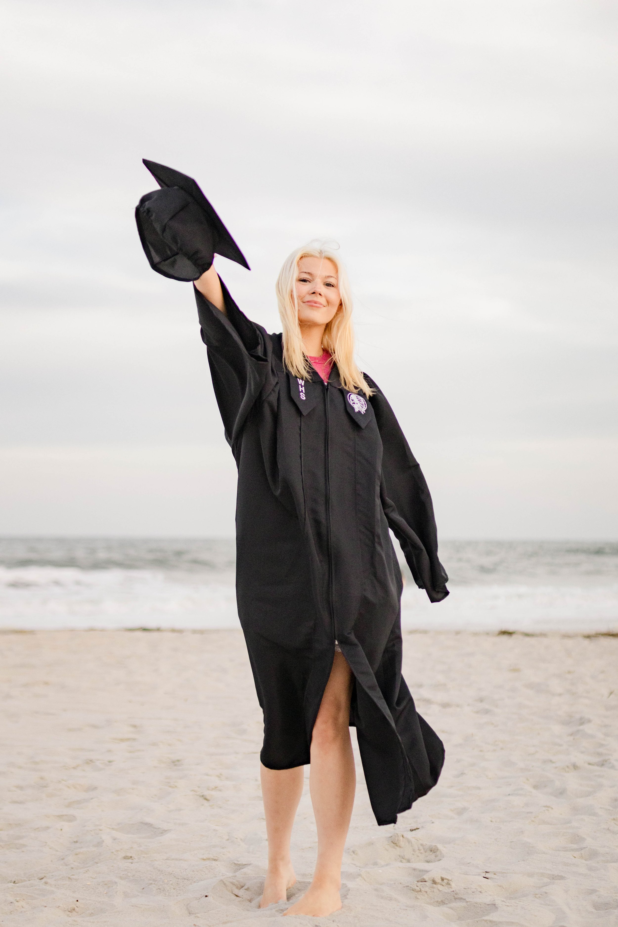 High school senior in Myrtle Beach wearing cap and gown during golden hour beach photo session, splashing in the water and smiling, taken by Myrtle Beach family and senior photographer.