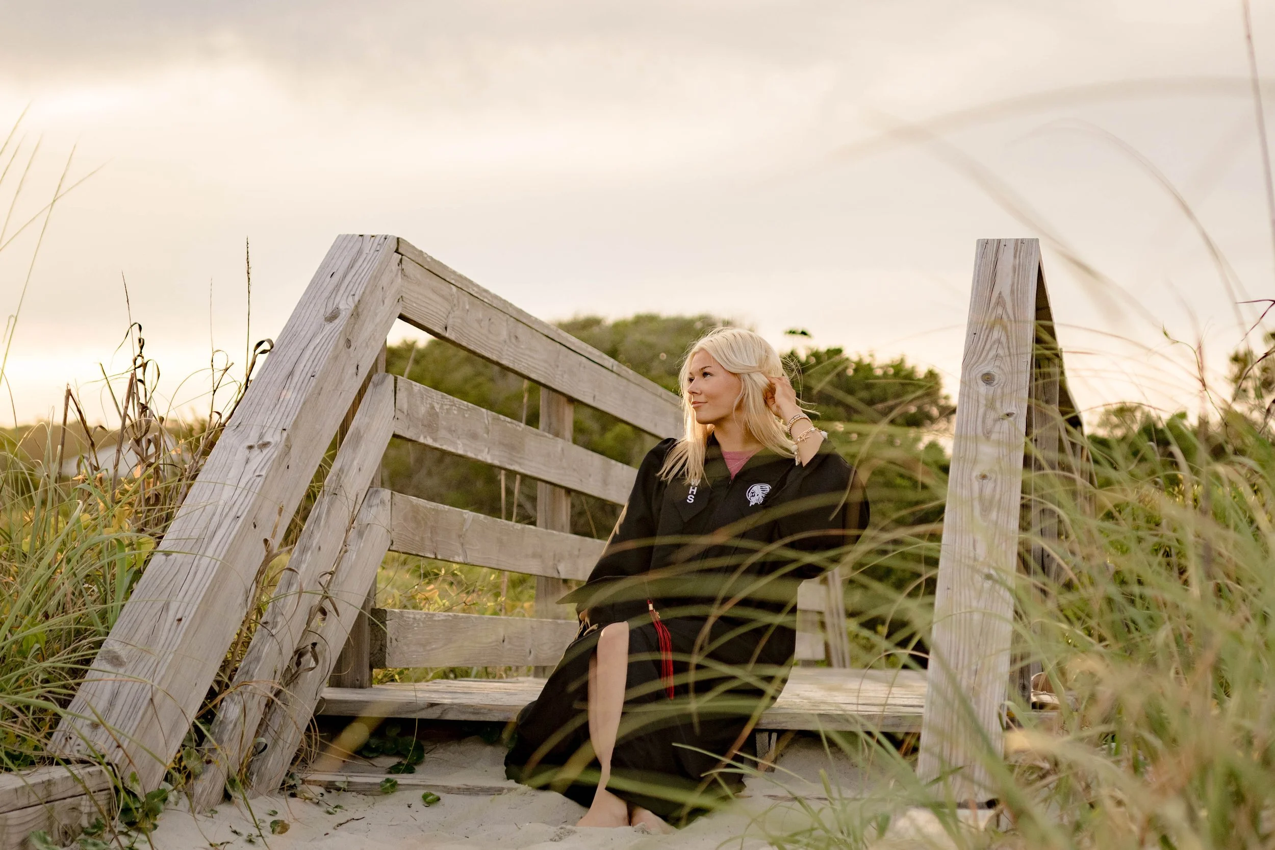 High school senior in Myrtle Beach wearing cap and gown during golden hour beach photo session, splashing in the water and smiling, taken by Myrtle Beach family and senior photographer.