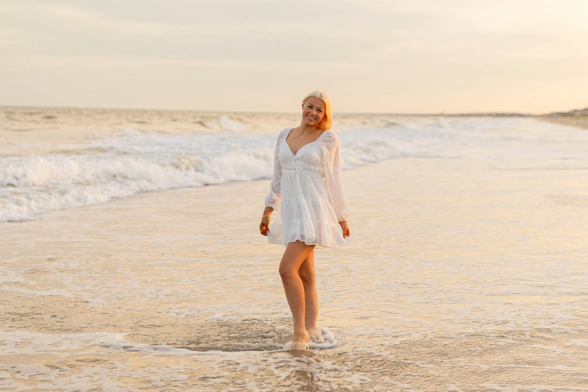 High school senior in Myrtle Beach wearing cap and gown during golden hour beach photo session, splashing in the water and smiling, taken by Myrtle Beach family and senior photographer.