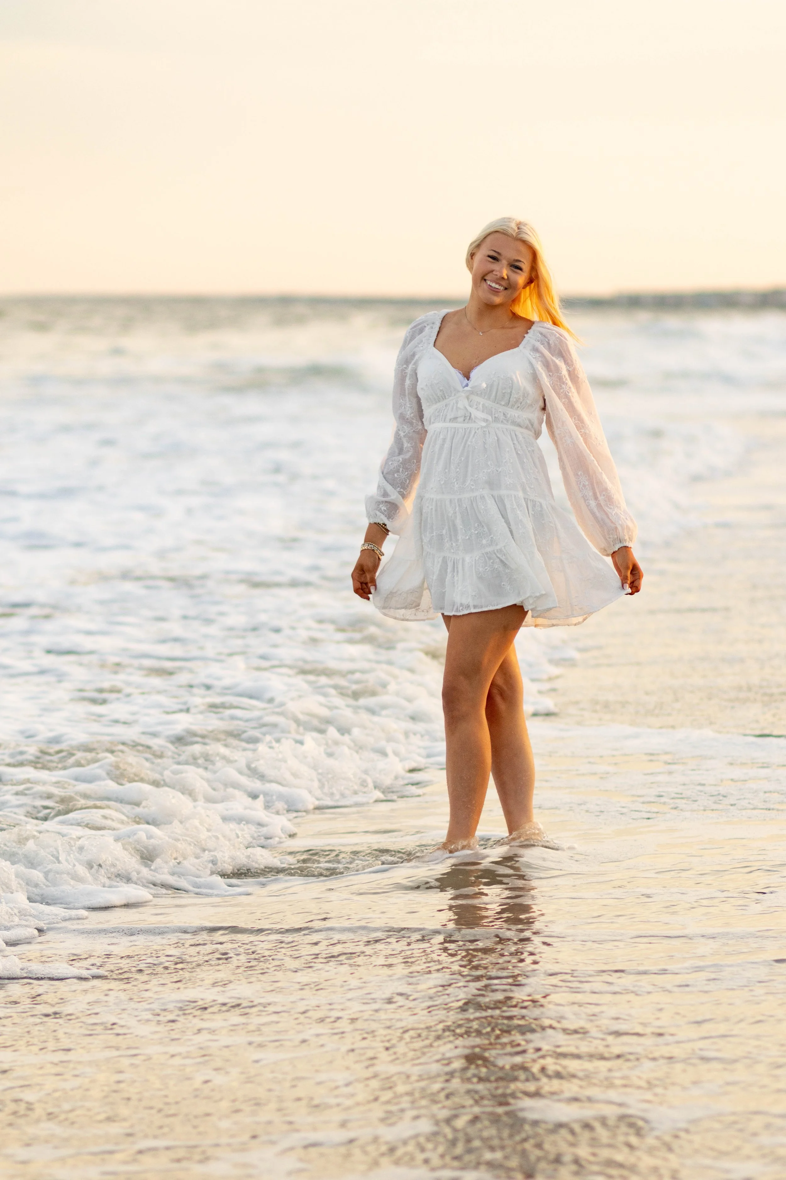 High school senior in Myrtle Beach wearing cap and gown during golden hour beach photo session, splashing in the water and smiling, taken by Myrtle Beach family and senior photographer.