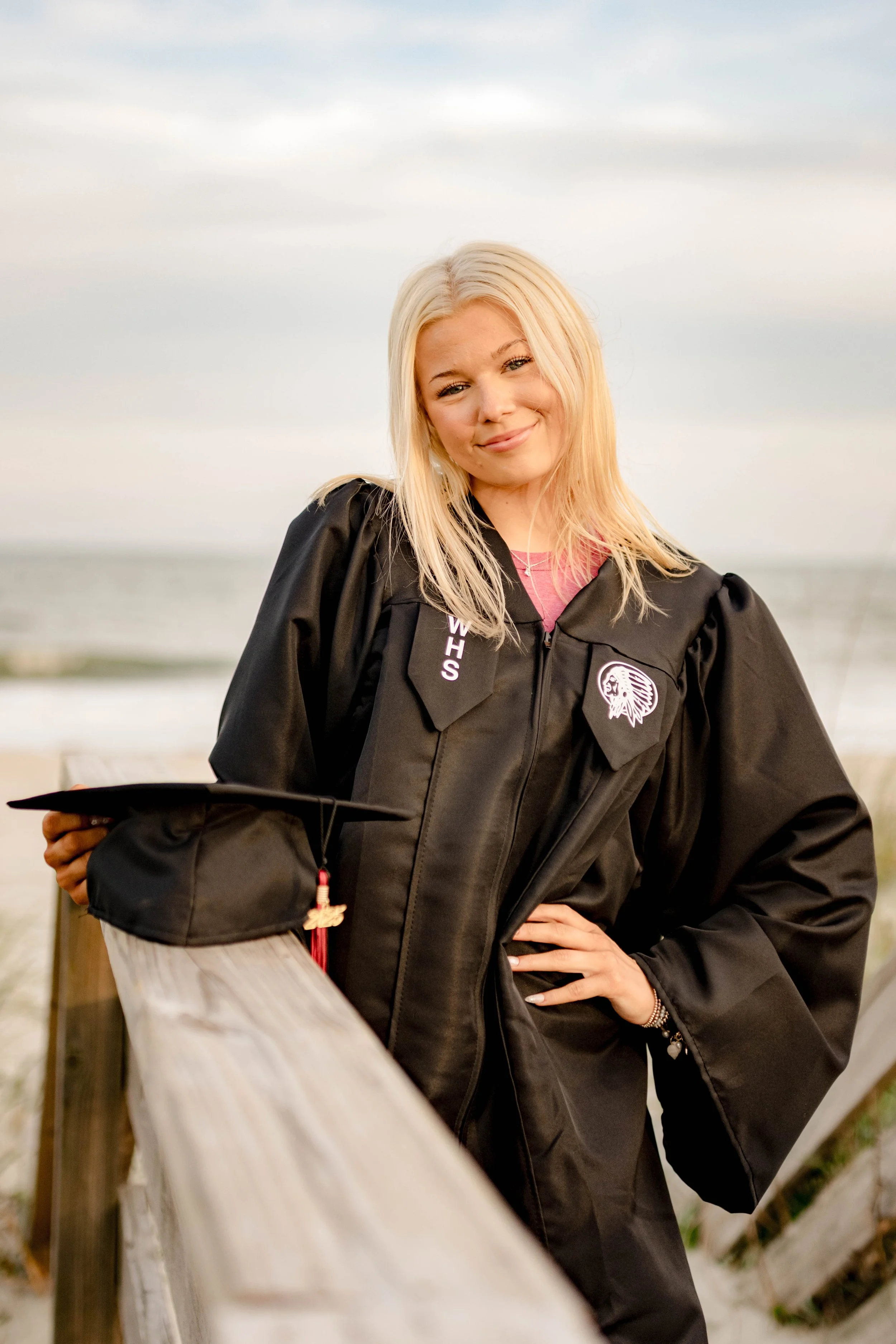 High school senior in Myrtle Beach wearing cap and gown during golden hour beach photo session, splashing in the water and smiling, taken by Myrtle Beach family and senior photographer.