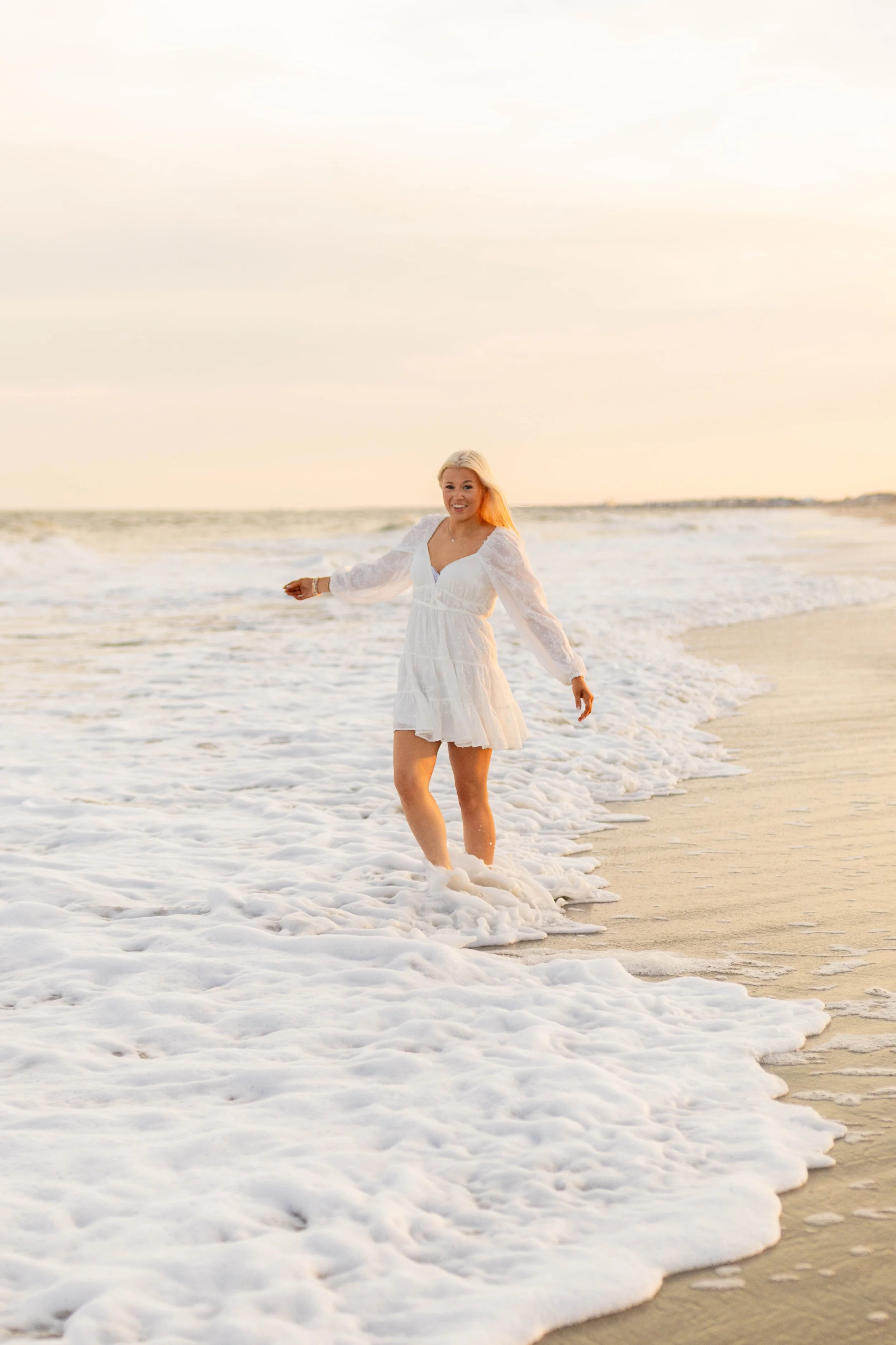 High school senior in Myrtle Beach wearing cap and gown during golden hour beach photo session, splashing in the water and smiling, taken by Myrtle Beach family and senior photographer.