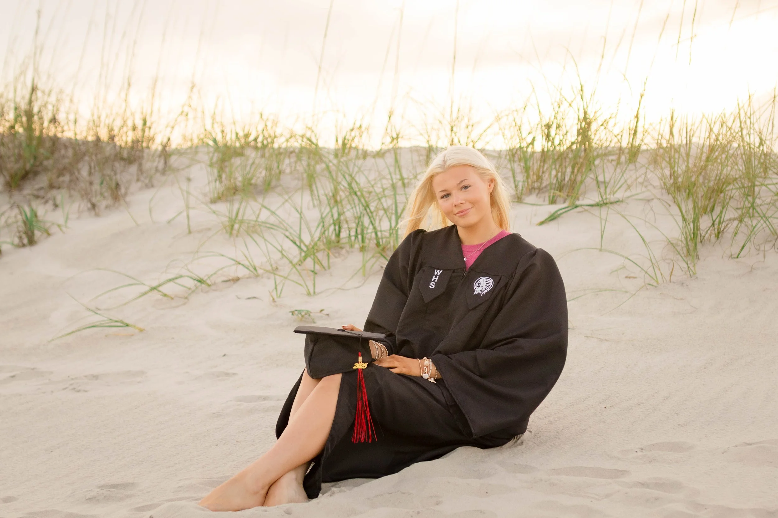 High school senior in Myrtle Beach wearing cap and gown during golden hour beach photo session, splashing in the water and smiling, taken by Myrtle Beach family and senior photographer.