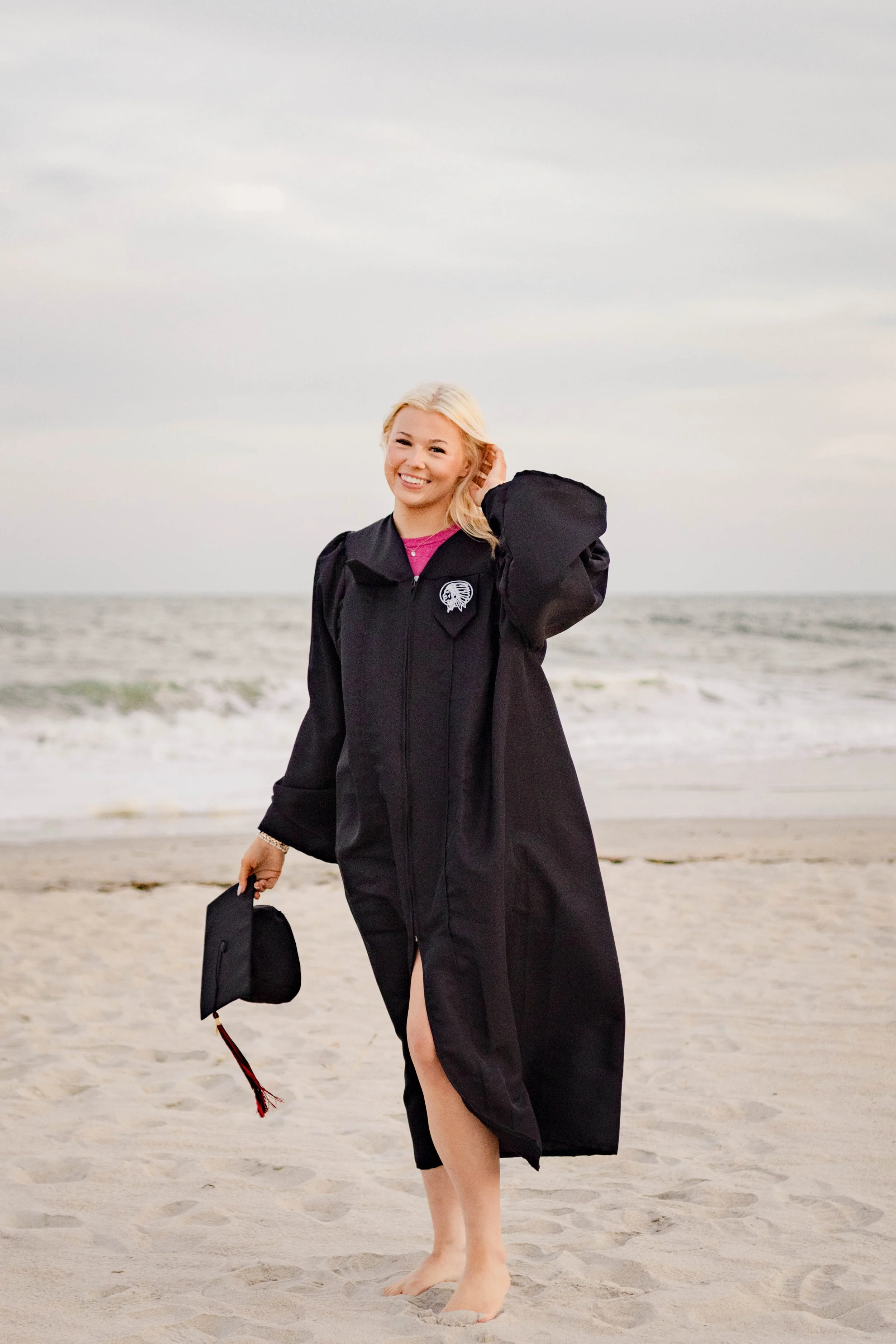 High school senior in Myrtle Beach wearing cap and gown during golden hour beach photo session, splashing in the water and smiling, taken by Myrtle Beach family and senior photographer.