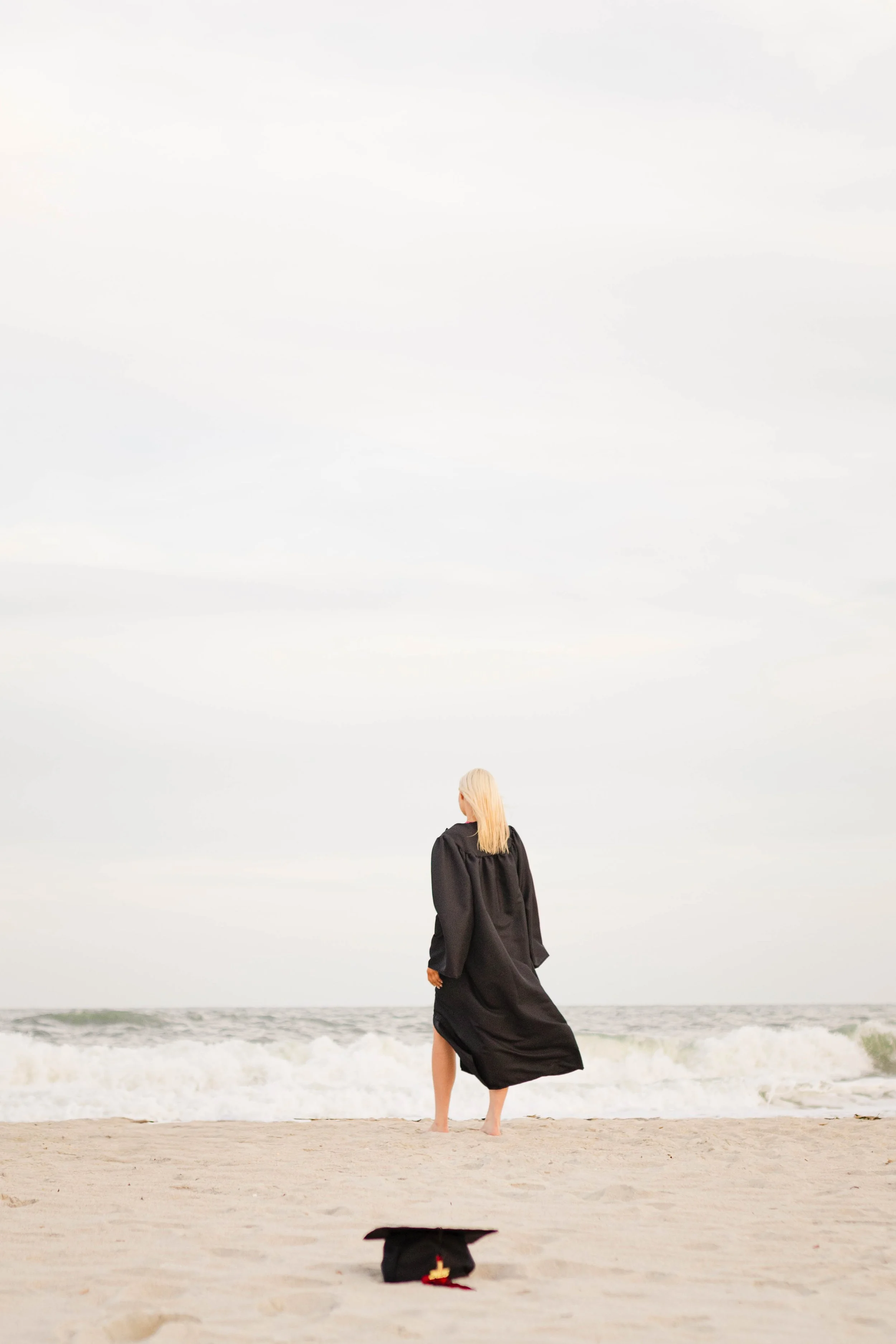 High school senior in Myrtle Beach wearing cap and gown during golden hour beach photo session, splashing in the water and smiling, taken by Myrtle Beach family and senior photographer.
