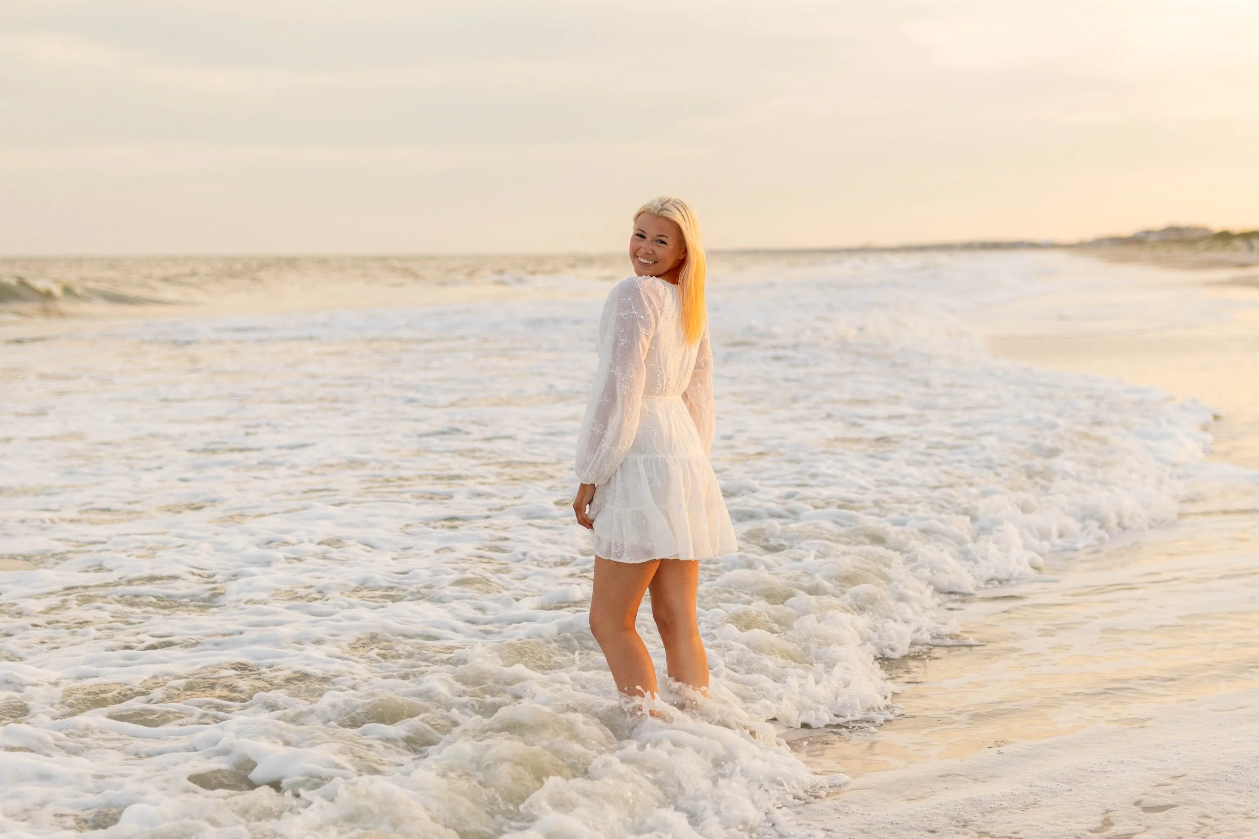 High school senior in Myrtle Beach wearing cap and gown during golden hour beach photo session, splashing in the water and smiling, taken by Myrtle Beach family and senior photographer.