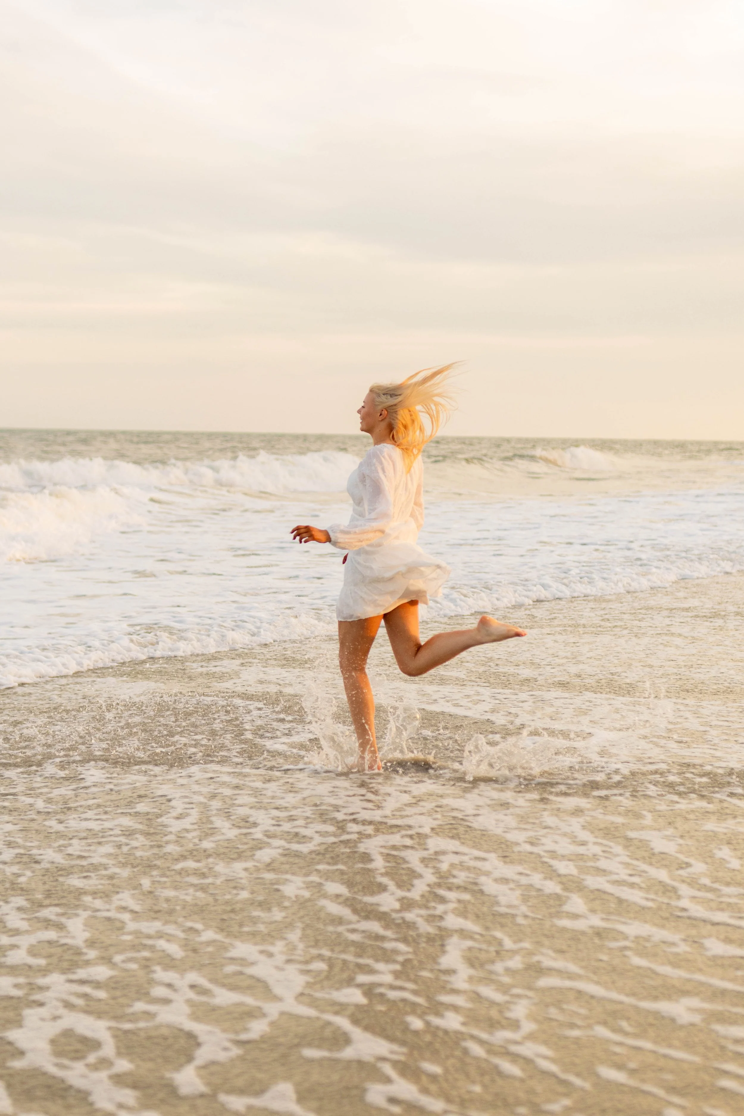 High school senior in Myrtle Beach wearing cap and gown during golden hour beach photo session, splashing in the water and smiling, taken by Myrtle Beach family and senior photographer.